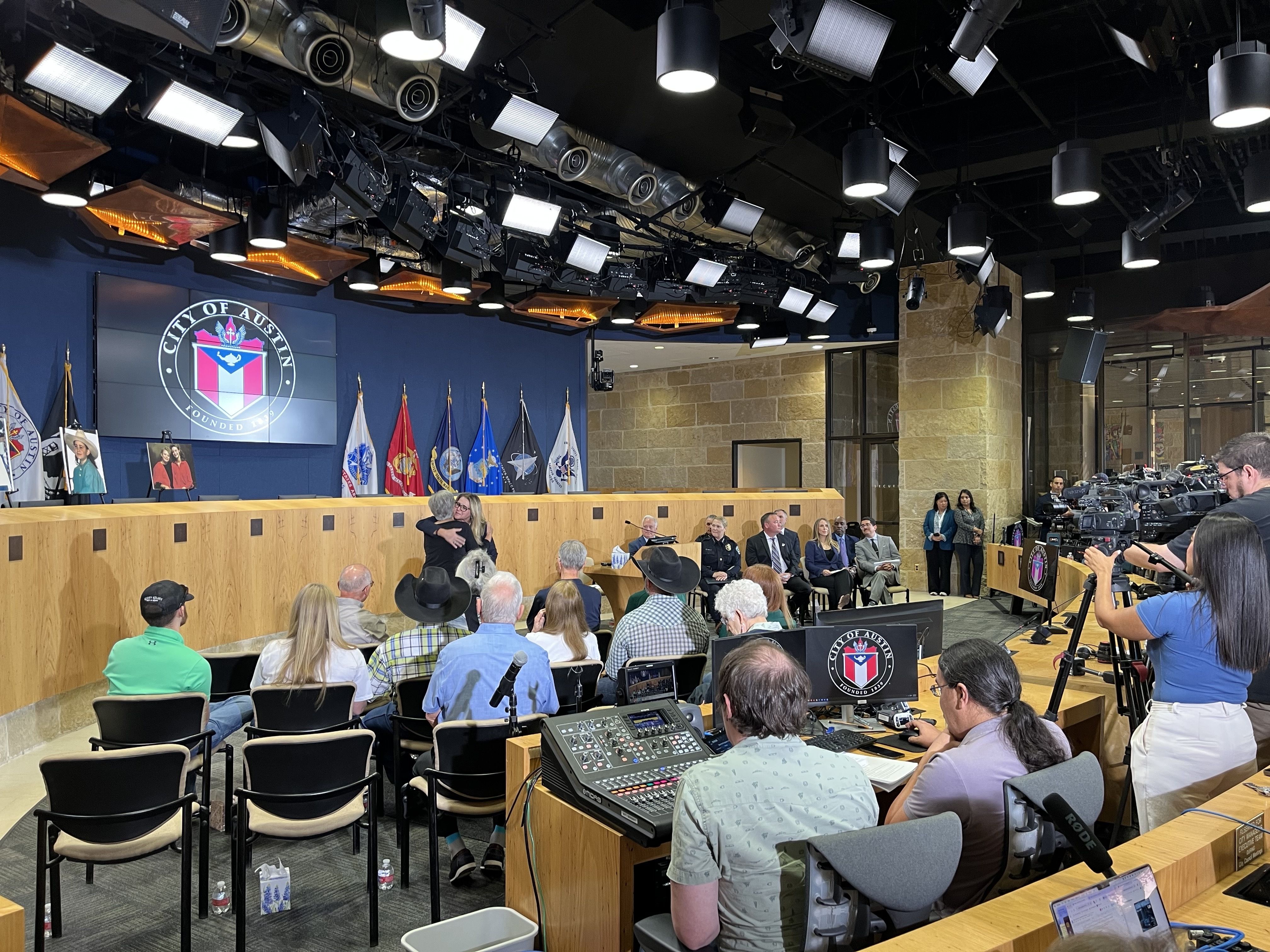 City of Austin council chamber with two people hugging center stage, flags and city emblem on screen, seated audience, officials at front, and media recording on the right.