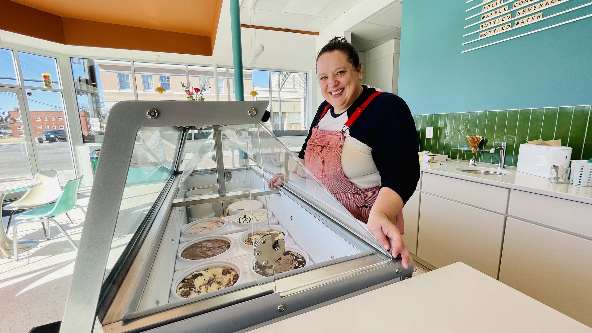 Smiling woman in pink overalls behind glass ice cream display with various flavors in bright, modern shop with green tiled wall and light blue paint.