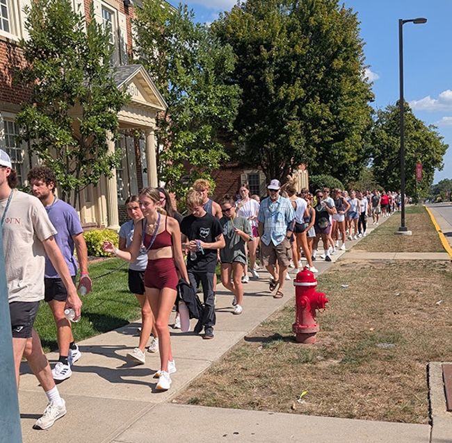 A line of college-aged people walk down a sidewalk on a sunny day. To their left is campus housing on a tree-lined street.
