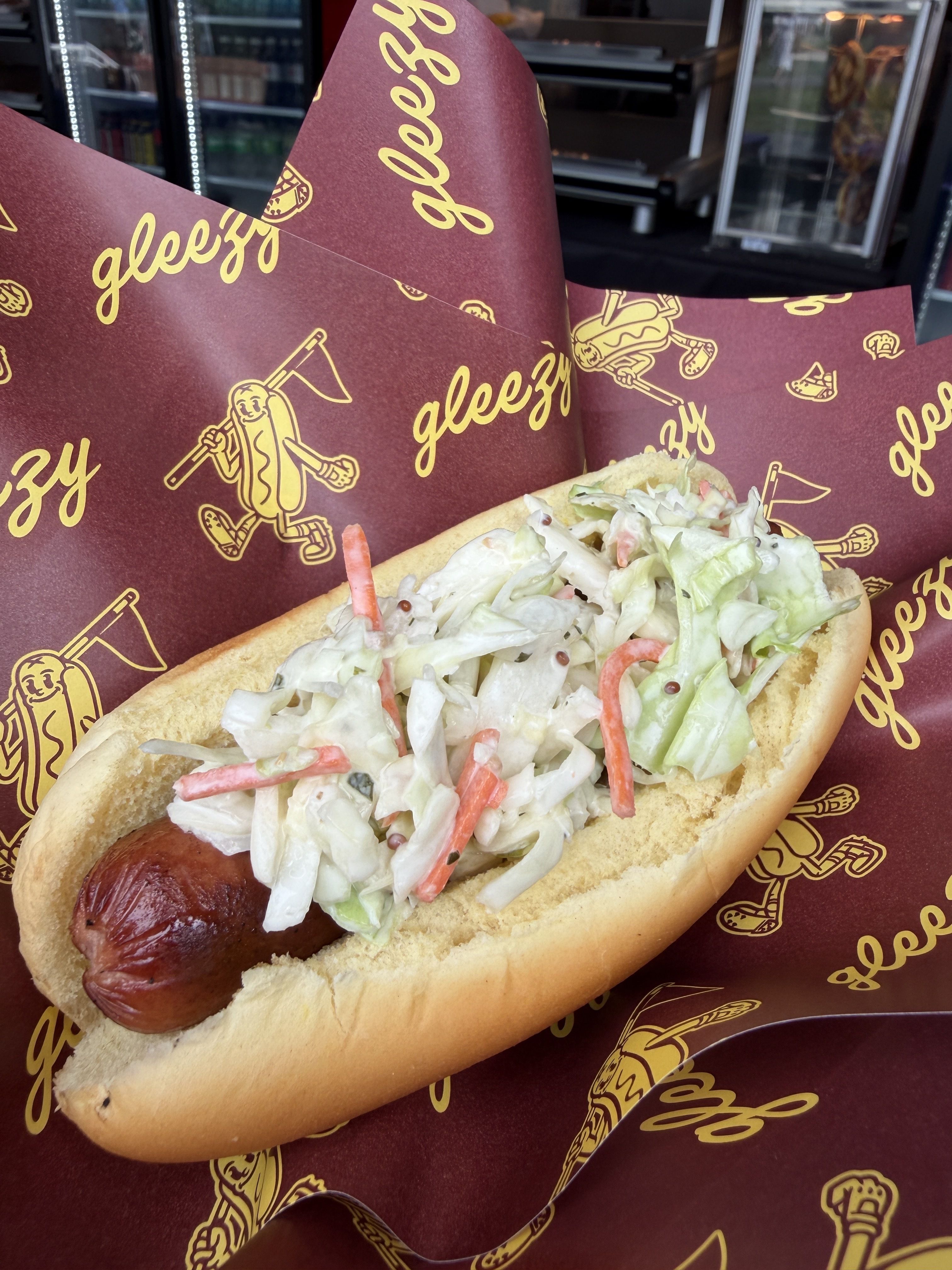 Hot dog in a bun topped with coleslaw and shredded vegetables, served on maroon wrapping with yellow "gleezy" logos; a fridge display is visible in the background.