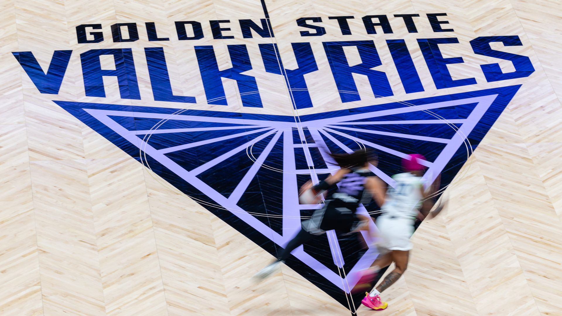 Photo of the Golden State Valkyries logo on a basketball court as two players vie for the ball