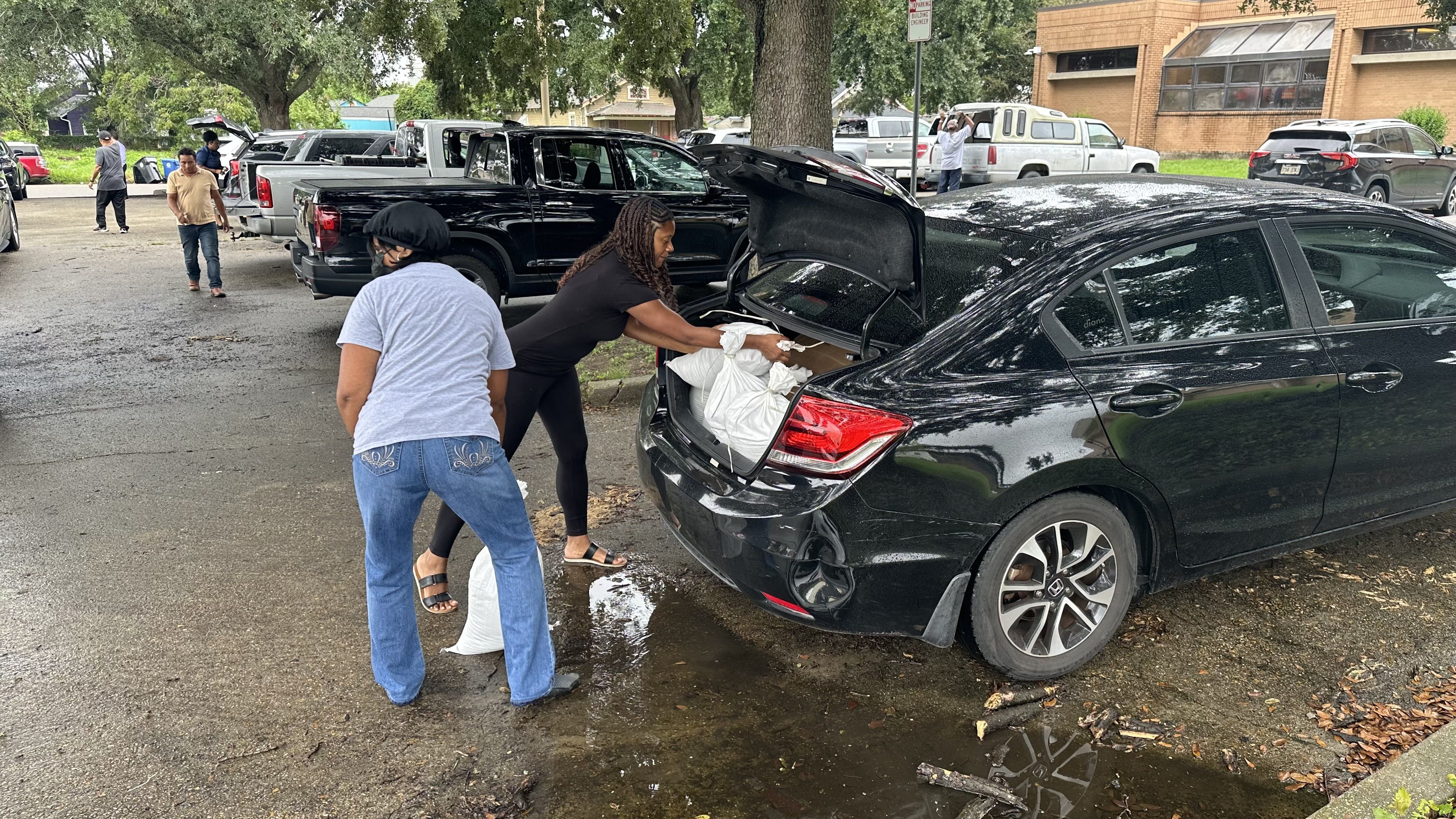 Two women load sandbags into the bag of a black sedan.