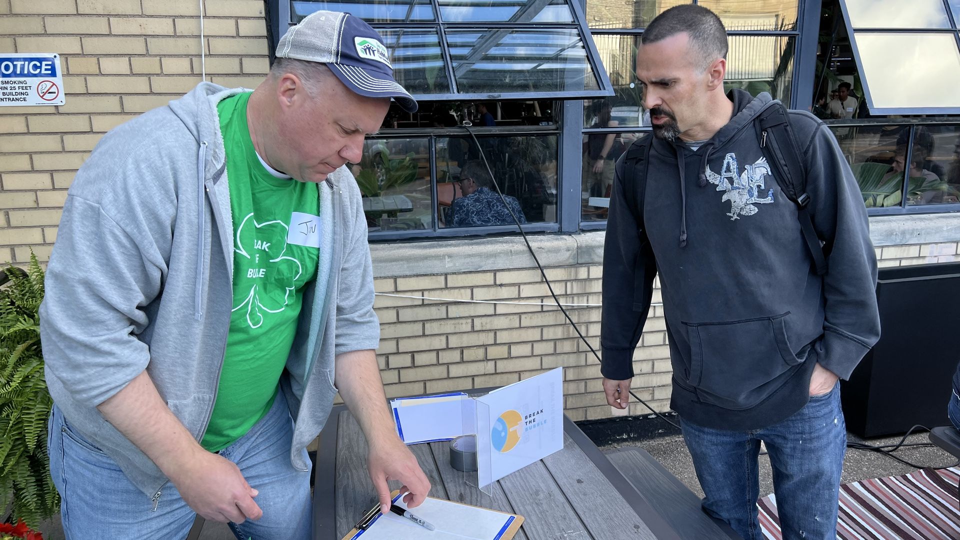 Two men, one in an unzipped hoodie and green t-shirt, the other in a dark blue hoodie, talk over a picnic table with a sheet of name tags on it and a sign that reads "Break the Bubble."