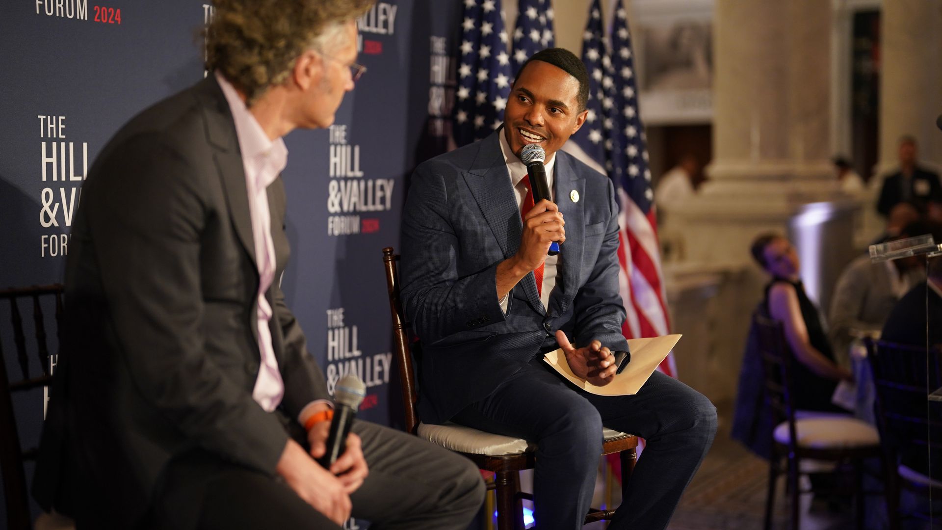 Rep. Ritchie Torres, wearing a gray suit, white shirt and red tie, speaking into a microphone at an event.