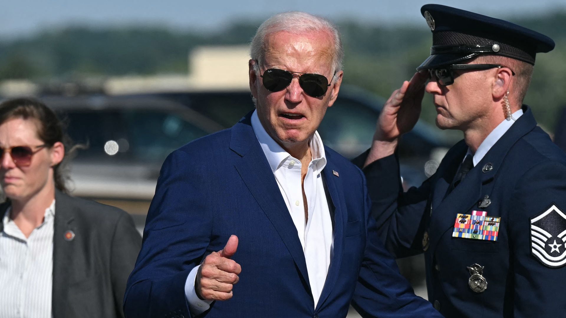 President Biden, wearing a blue suit and giving a thumbs up flanked by a soldier and a member of his security detail on a tarmac.