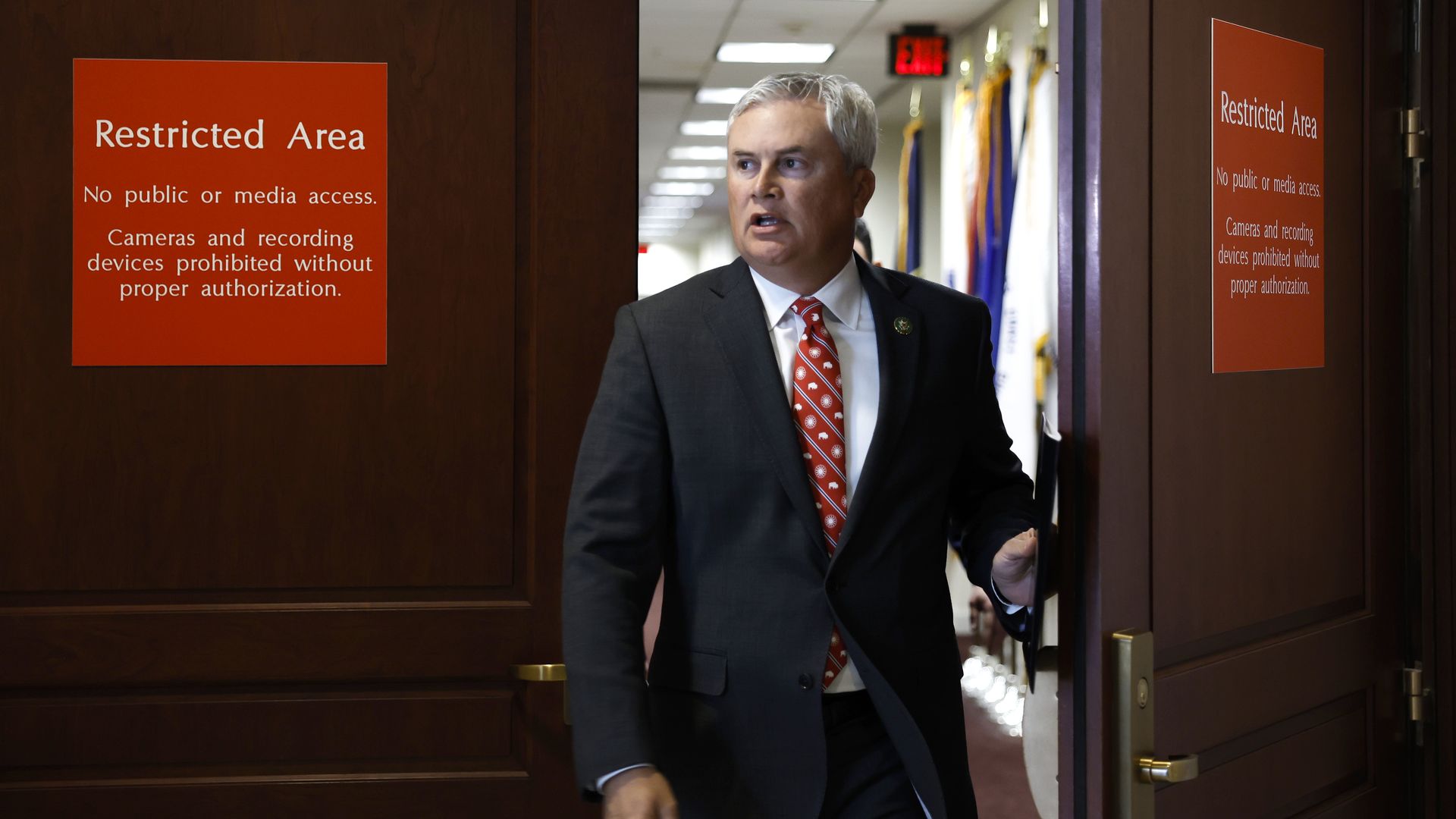 Rep. James Comer, wearing a gray suit and walking through a wooden door with red signs that say "restricted area."