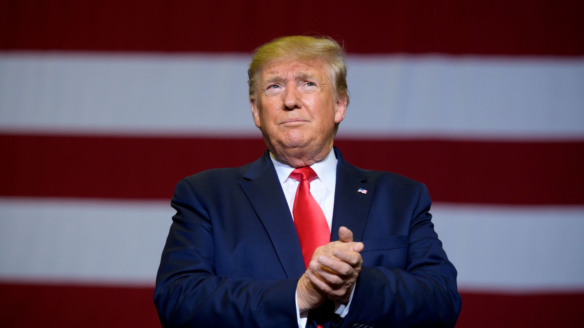 In this image, Trump stands in front of a large American flag with his hands clasped together.