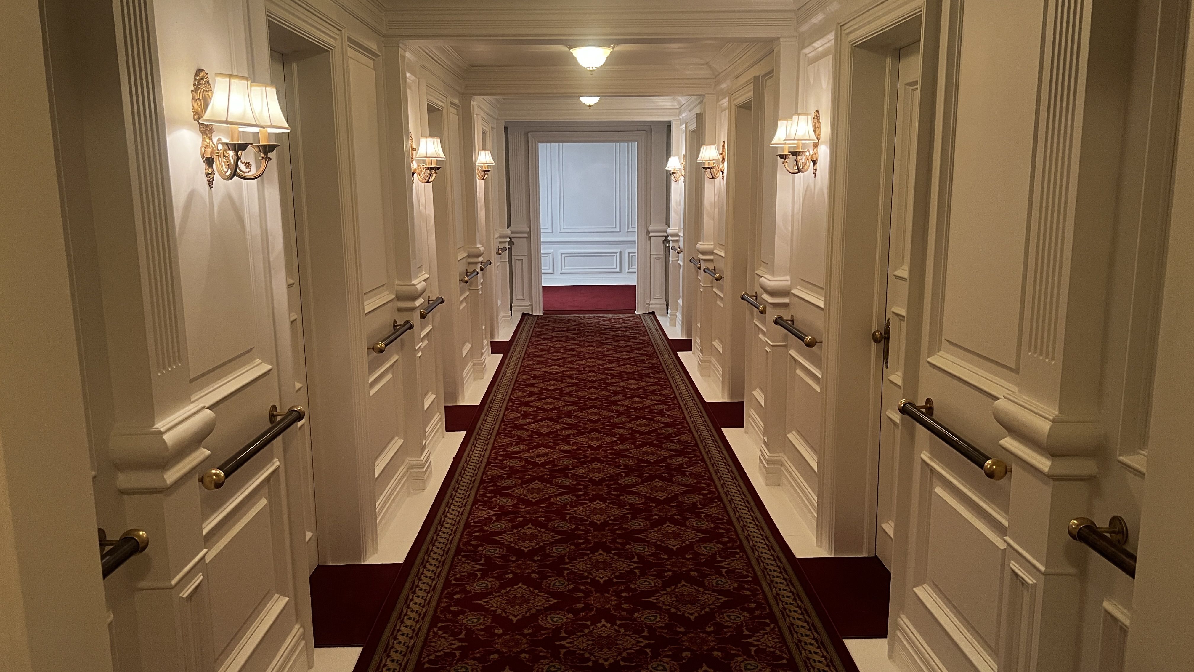 A long, upscale hotel hallway with white paneled walls, ornate wall sconces, and a burgundy patterned runner carpet stretching toward a distant doorway.