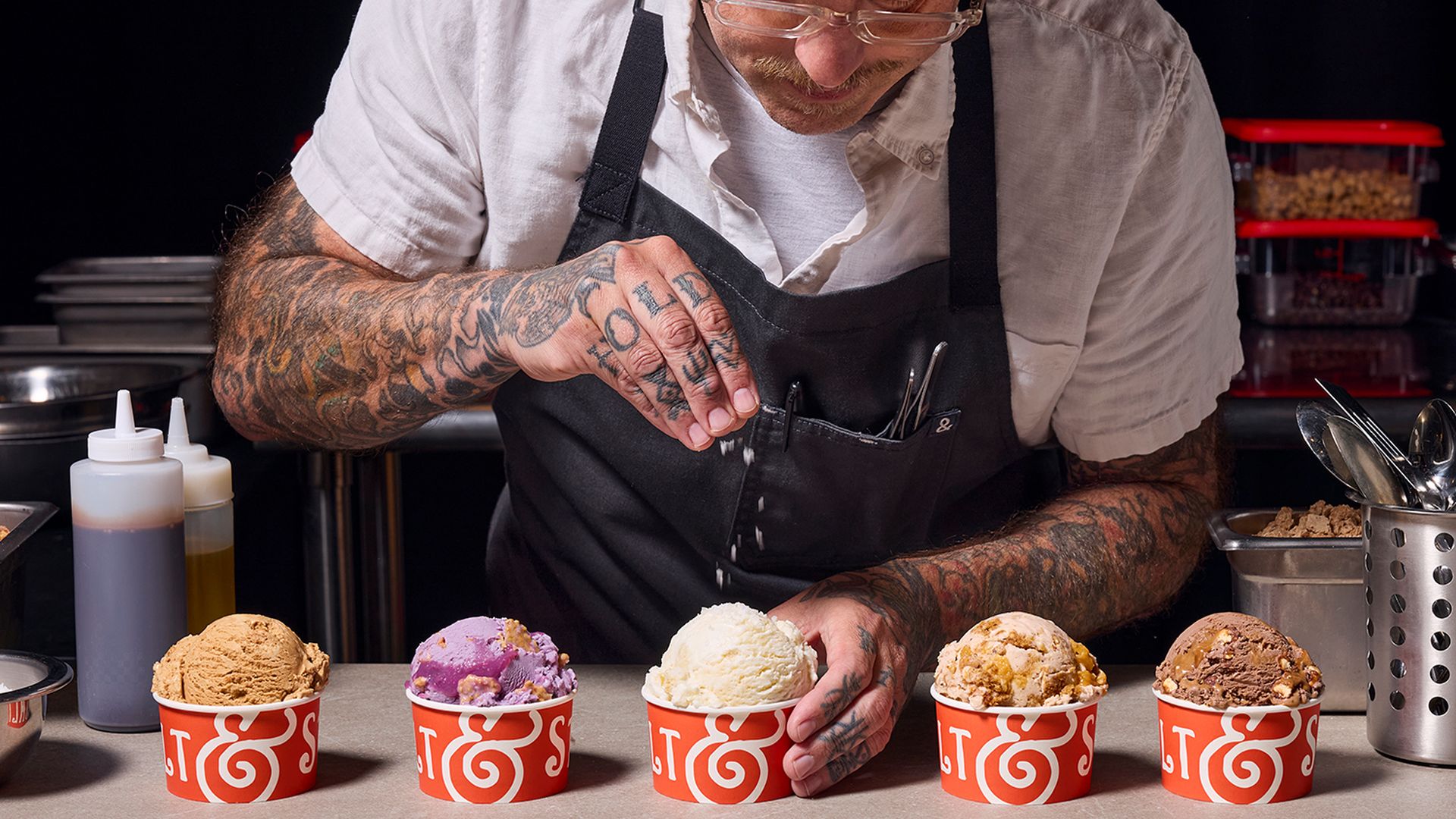 Tattooed chef in a white shirt and black apron sprinkles salt on five ice cream scoops in red cups, with a "THE CHEF SERIES" banner and Salt & Straw logo visible.