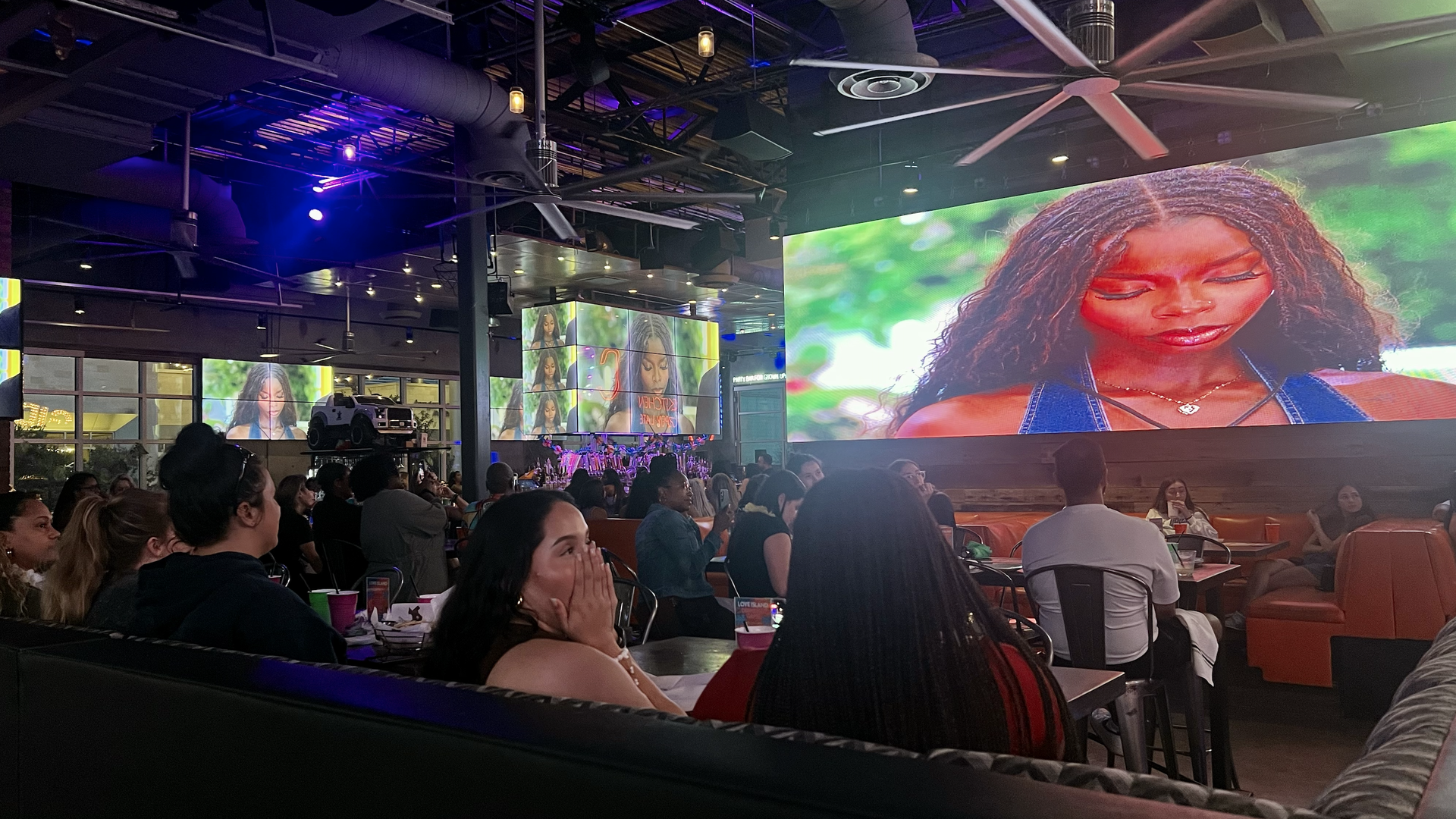 Dozens of people sit in a bar while TV screens show a woman looking down