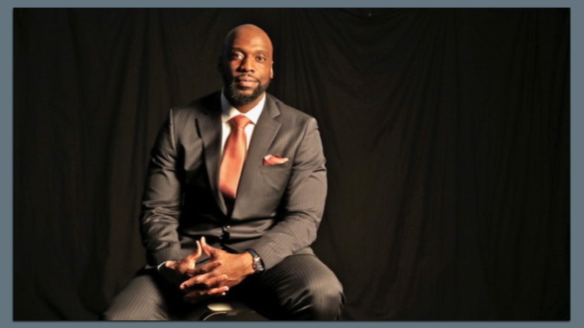 Former NFL player and current therapist Tim Massaquoi wearing a dark pinstripe suit with a white shirt and orange tie, sitting with hands clasped, against a dark background.