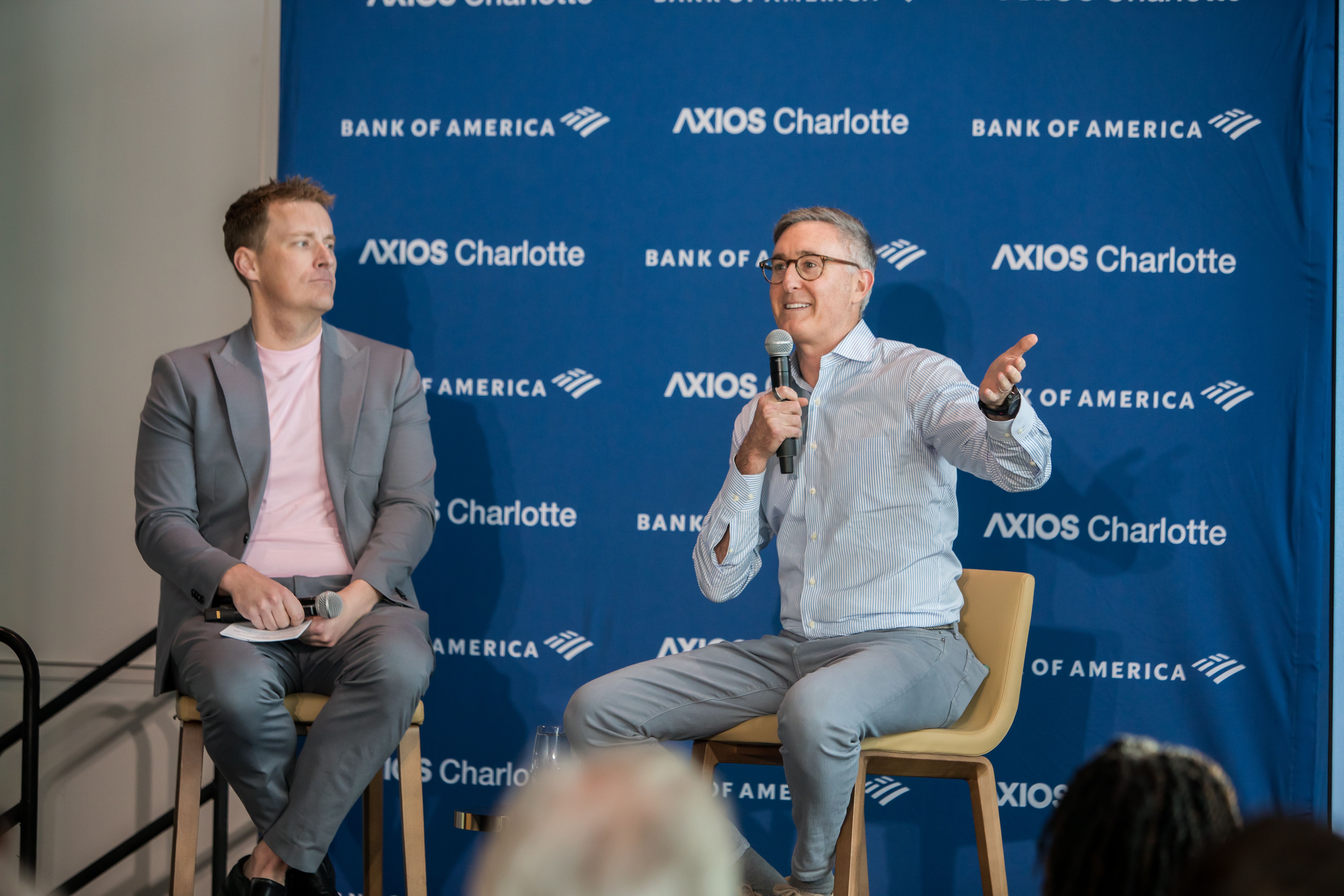 Two men sit on a stage in front of a blue backdrop with AXIOS Charlotte and Bank of America logos. Left: gray suit and pink shirt; right: glasses, striped shirt, speaking into a mic and gesturing.