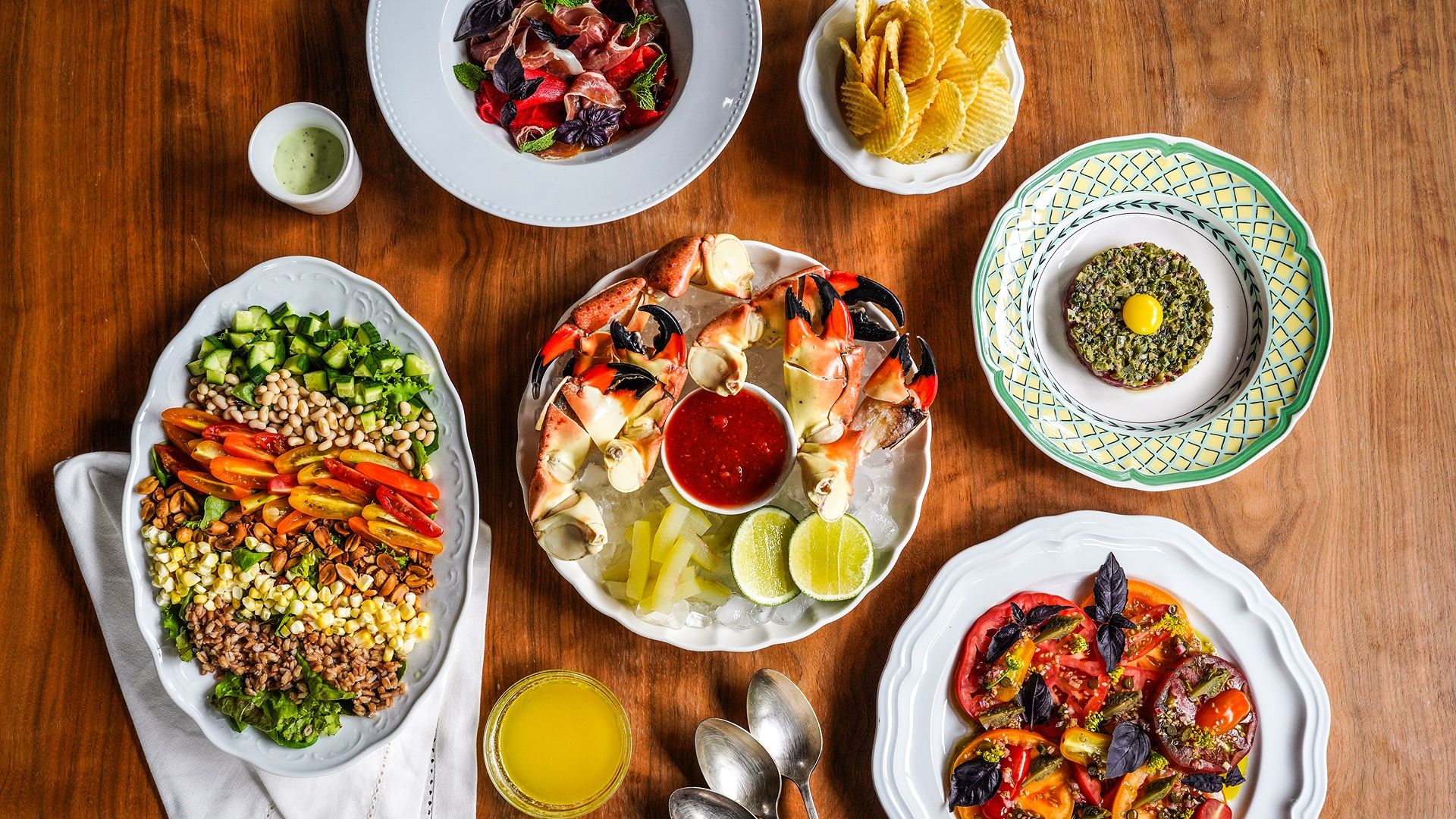 Overhead view of a wooden table with a colorful spread: center shrimp platter with dipping sauce, a plate of diced vegetables, chips, lime wedges, and various other plated dishes.