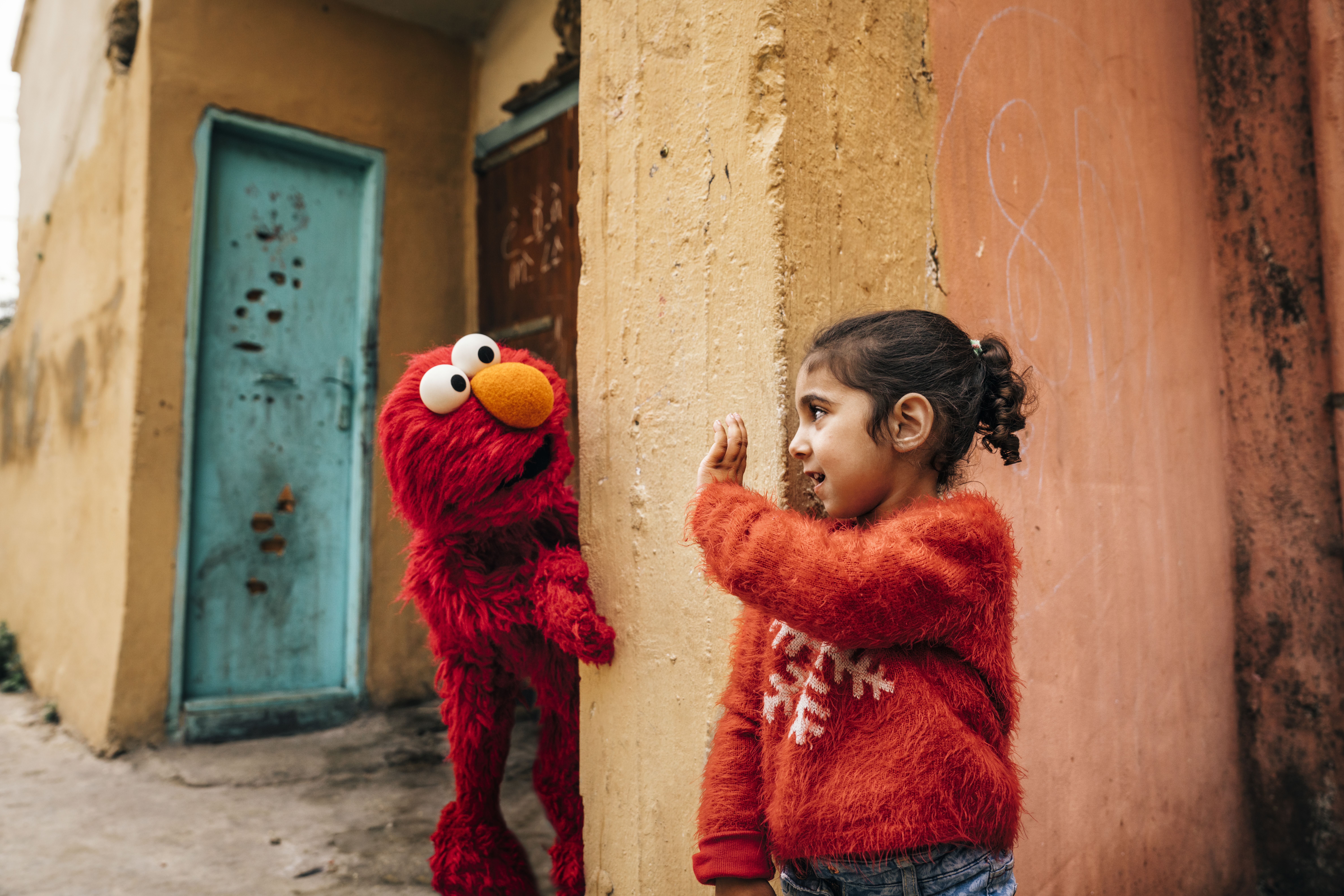 photo of Elmo, the muppet from Sesame Street, looking at a young girl who is standing on the corner of a colorful building
