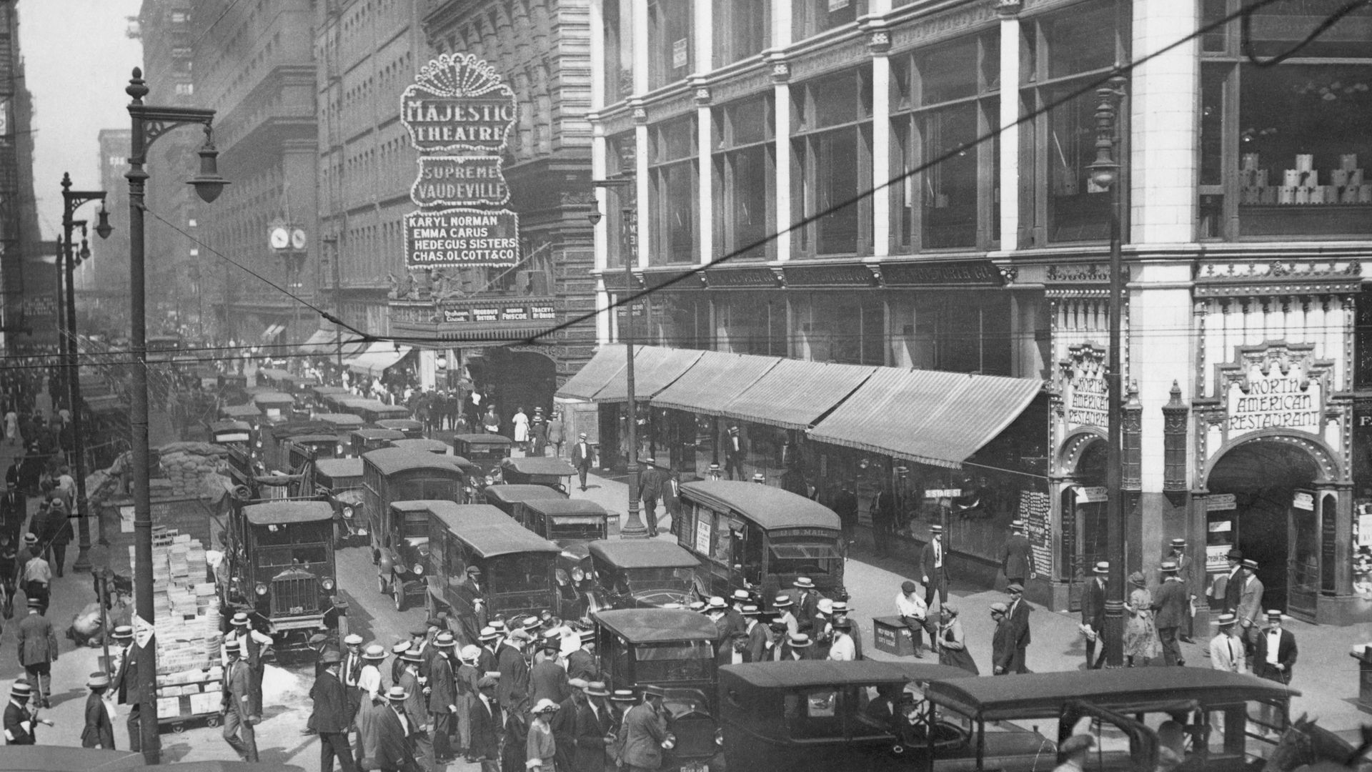 Black-and-white street scene from the early 20th century: crowded avenue with horse-drawn carriages, trolley wires, tall buildings, storefronts, and a Majestic Theatre marquee.