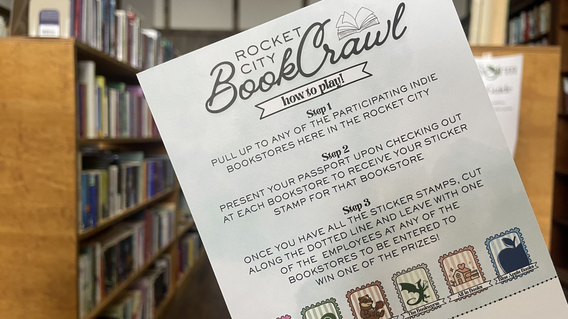 A tilted flyer in a library reads "Rocket City Book Crawl" with an open-book icon and "how to play!" text, set against shelves of books.