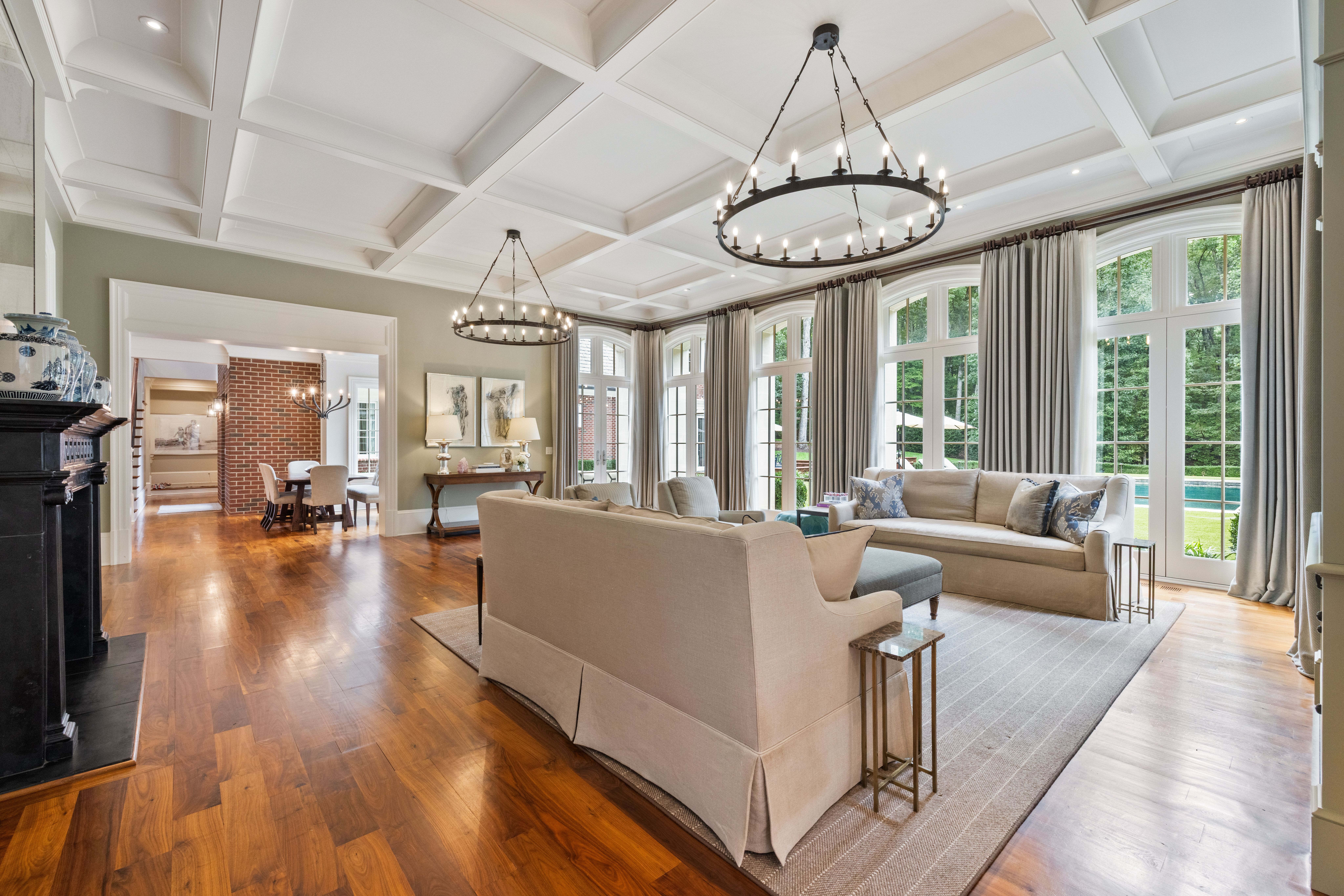 Bright living room with beige sofas, wooden floors, large tall windows with gray curtains, circular chandeliers, and a view of green trees and a pool outside.