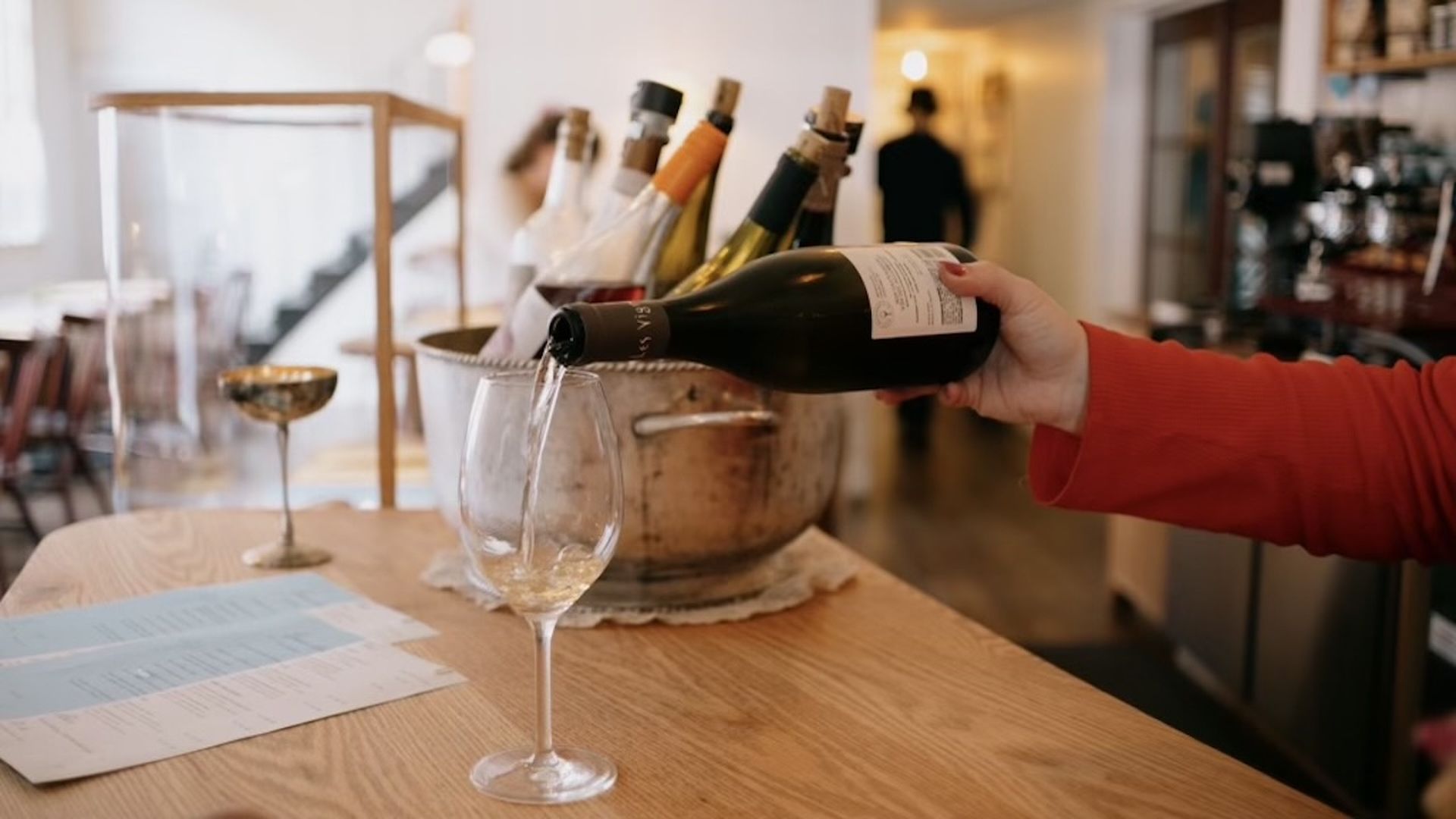 A photo of a person pouring a bottle of wine into a tall stem wine glass on a teak countertop. 