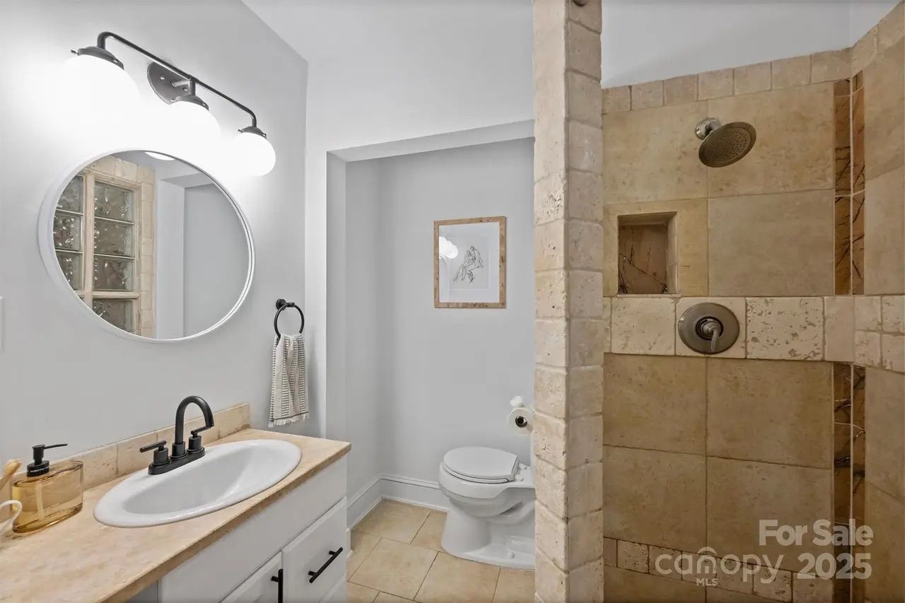Small bathroom with white walls, beige tile shower with bronze fixtures, white sink with beige countertop, round mirror, black faucet, and a toilet beneath a framed floral drawing.
