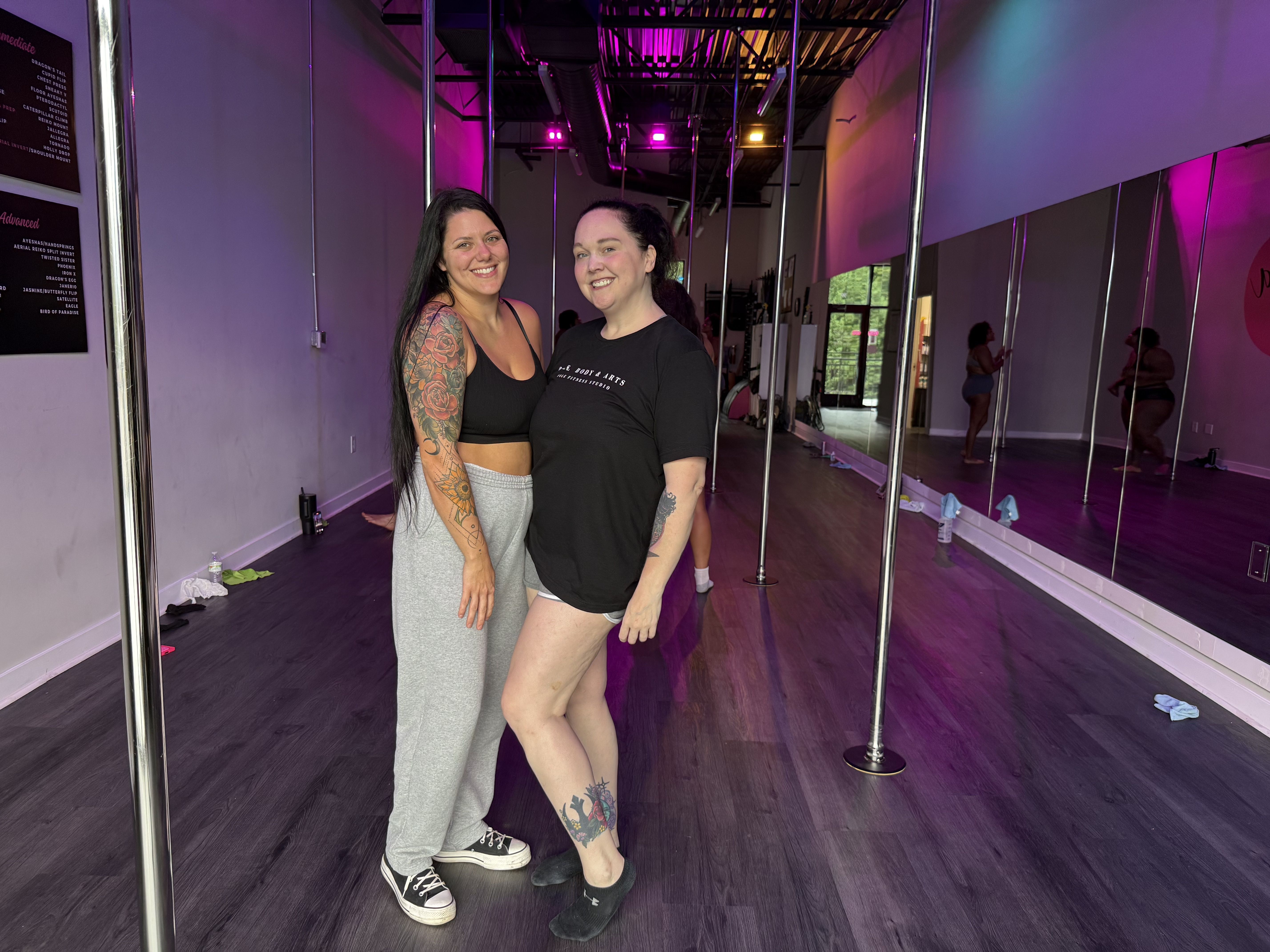 Two smiling women with tattoos pose inside a pole dancing studio with purple lighting, wooden floor, mirrors, and several poles. One wears black crop top and gray pants, the other black t-shirt and shorts.