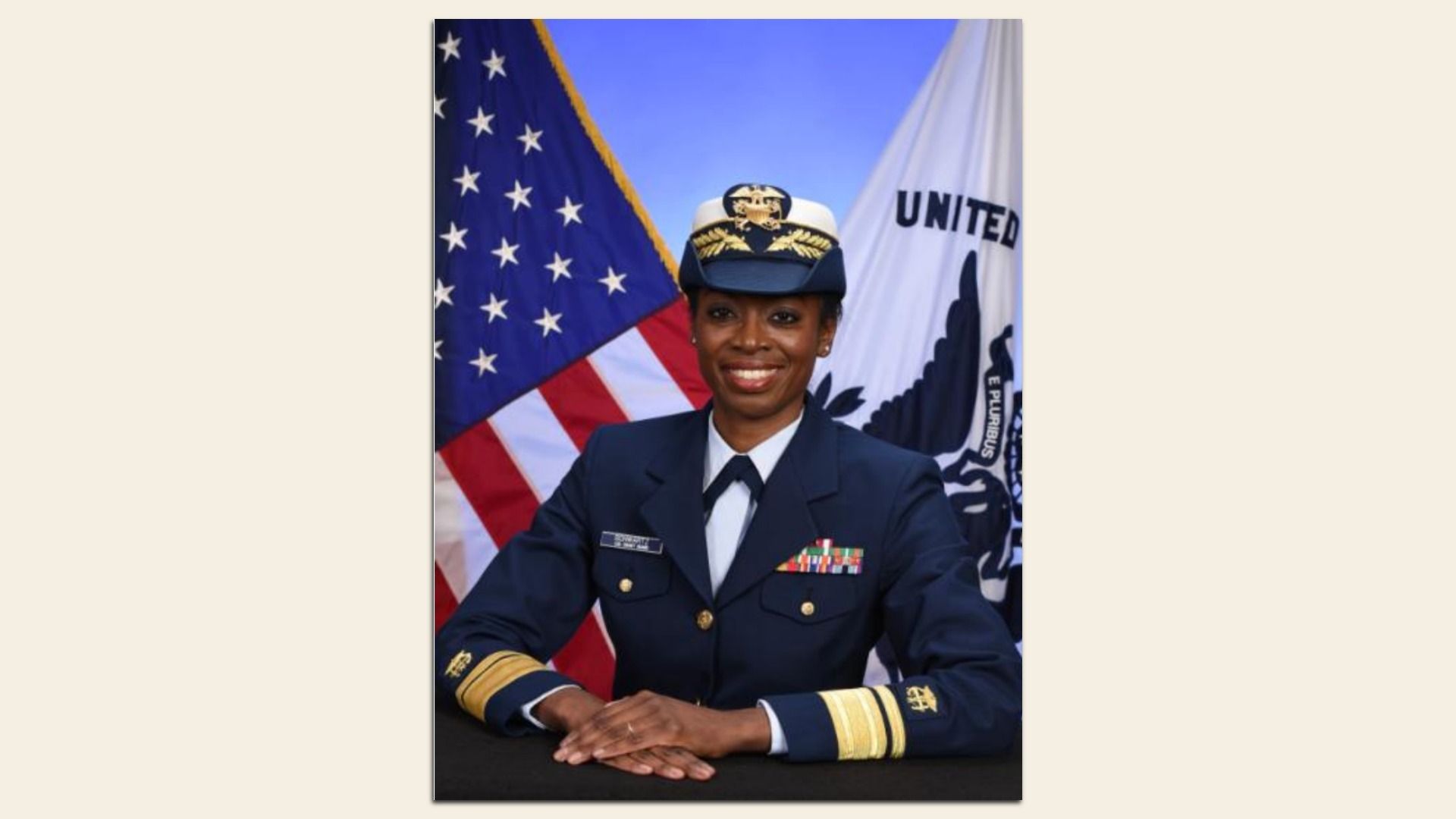 Portrait of Erica Schwartz in a navy dress uniform with ribbons, seated at a table with hands folded, in front of the U.S. flag and a white flag with an eagle.