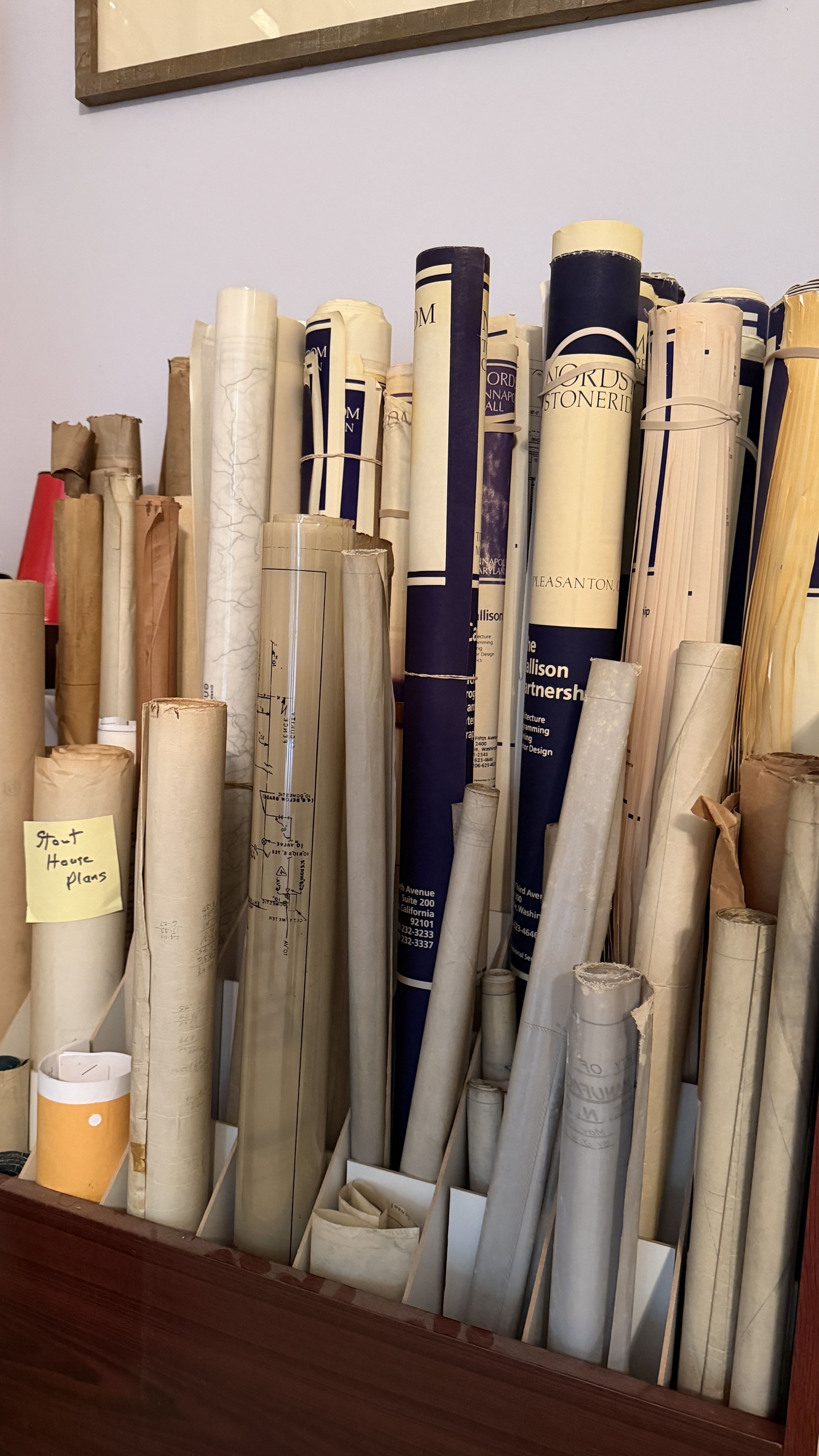 Rows of rolled architectural blueprints and drawings stand upright in a wooden desk rack. Beige and navy tubes fill the rack; a yellow sticky note reads "Stout House Plans".