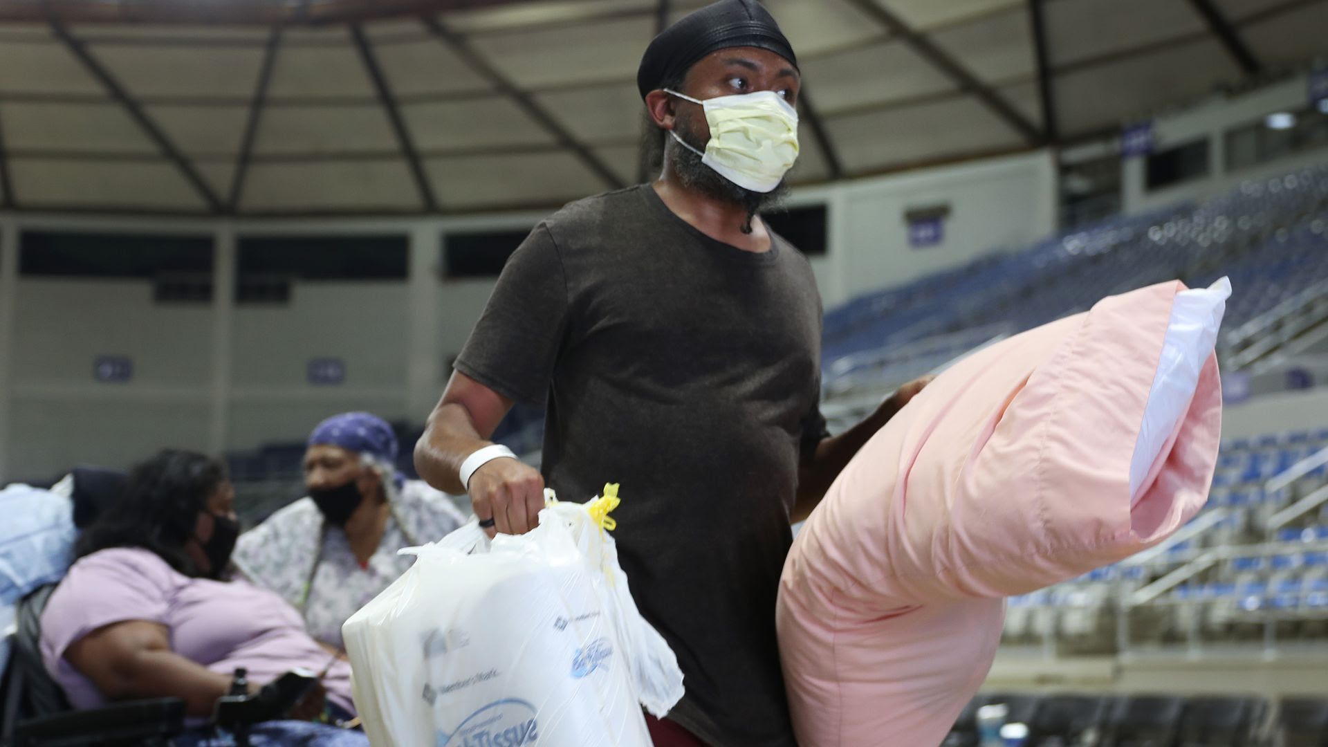 A man wering a mask carries a bag full of supplies and a pillow at a hurricane shelter