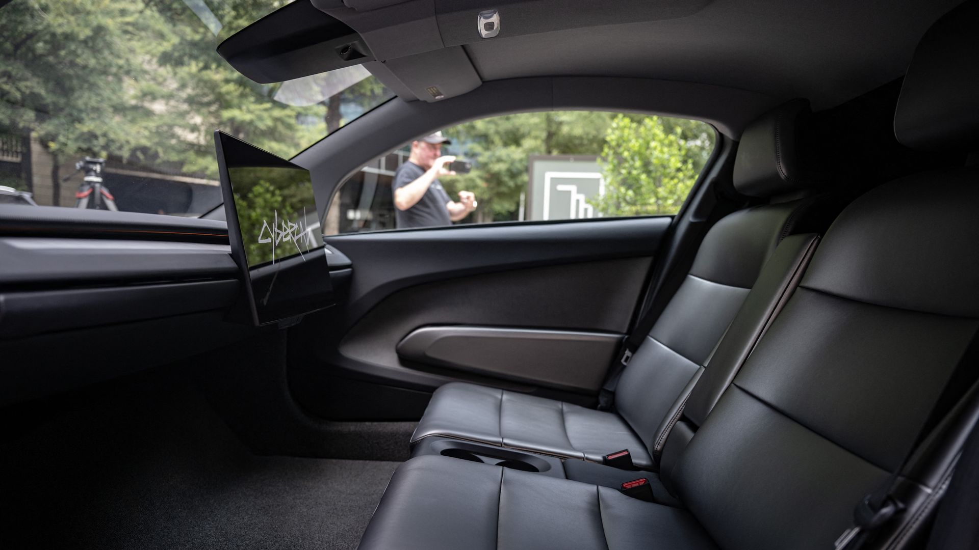 Interior of a dark car with black leather seats; a wide screen on the dashboard shows Cybercab white lettering. Through the window, a man in a cap takes a photo outside, with greenery and a tripod visible.