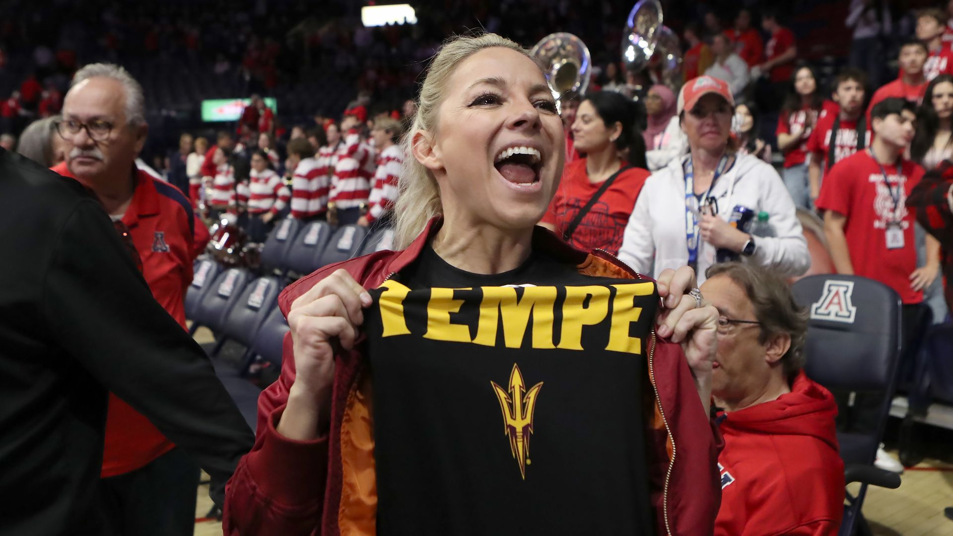 A woman with blond hair wearing a red jacket over a black shirt with the word "Tempe" and a pitchfork in yellow, yells cheerfully while pulling on the part of her shirt that says "Tempe." People stand and sit in arena seating behind her. 