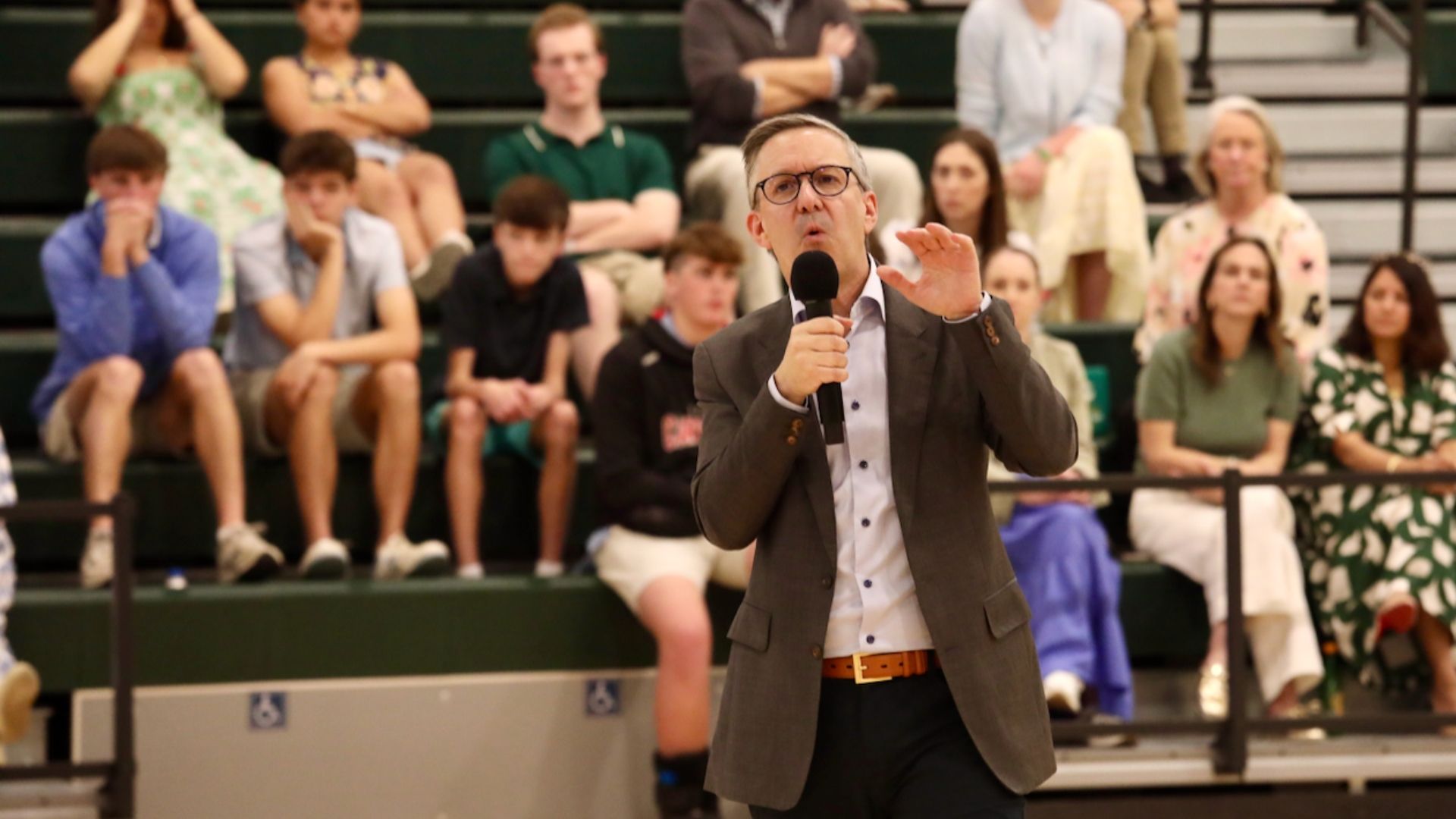Man in a gray suit and glasses speaks into a handheld microphone on a gym bleacher, gesturing with his left hand. Behind him, spectators sit on green bleachers.
