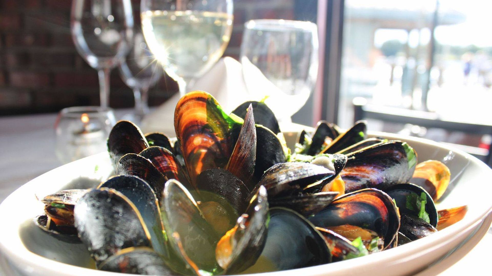 Close-up of a white bowl filled with cooked mussels in a light sauce, garnished with greens, on a table with wine glasses and blurred background of a restaurant by a window.