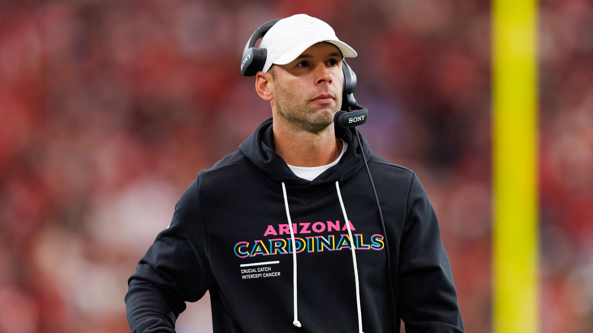 A man in a black hooded sweatshirt with "Arizona Cardinals" on the front, wearing a headset over a white ballcap, looks ahead on a football field.