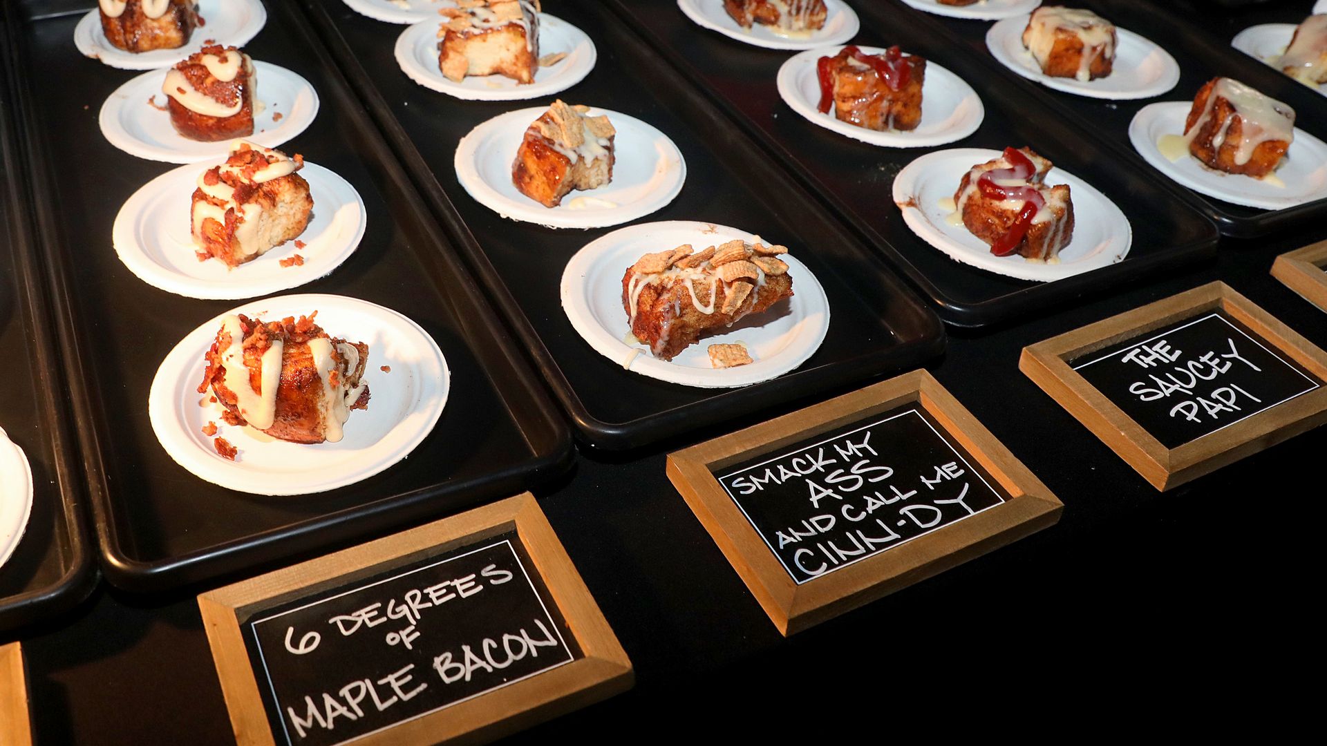 Display of slices of cinnamon bread on white plates arranged on black trays, each drizzled with icing and toppings; chalkboard signs indicate flavors such as Maple Bacon.