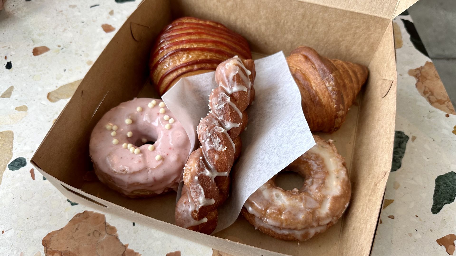 A box of pastries including two doughnuts, a twist and two croissants, sitting on a flecked marble table. 