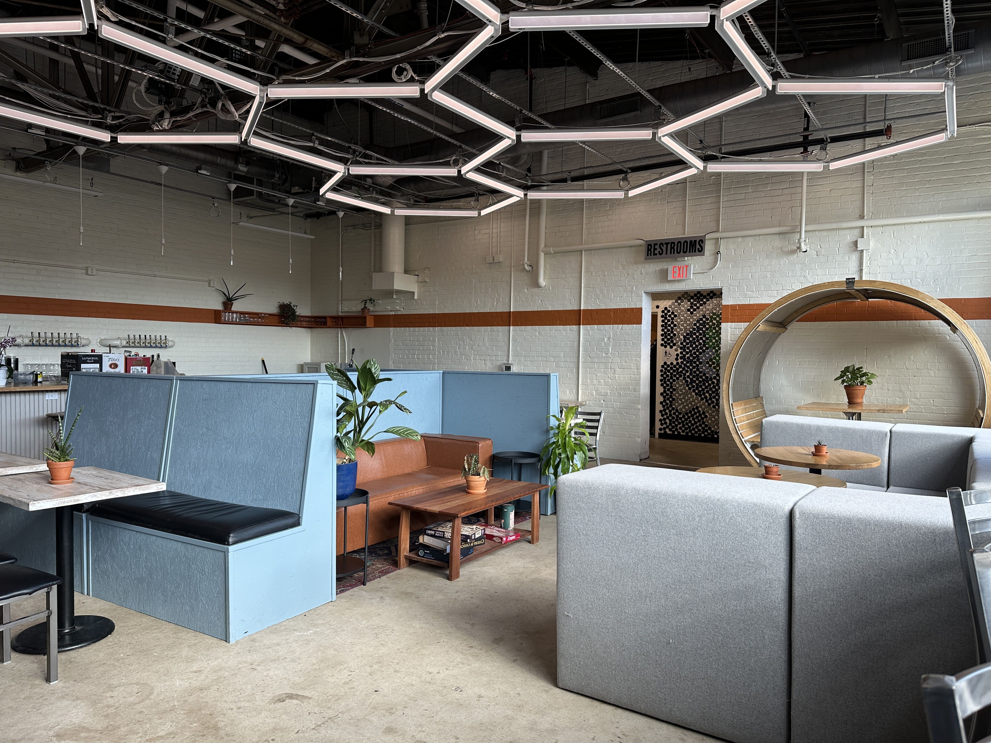 Modern cafe lounge with gray and brown seating, blue partitions, and potted plants. White hexagonal neon ceiling lights form a geometric grid. Brick walls with an orange stripe; restrooms visible. And/or cafe is a new concept from former Lorem Ipsum owner that opens in Belmont neighborhood in the former Change Please cafe space inside Innovation Barn.