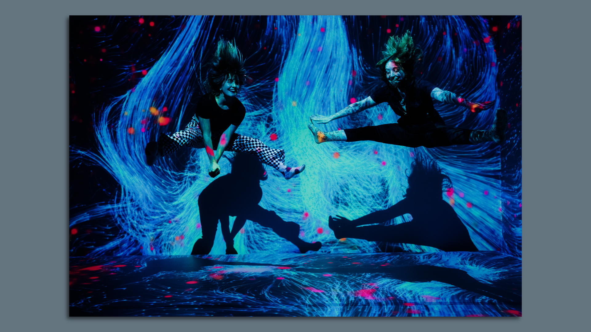 Two women bouncing on a trampoline doing the split in the air in a black lit room