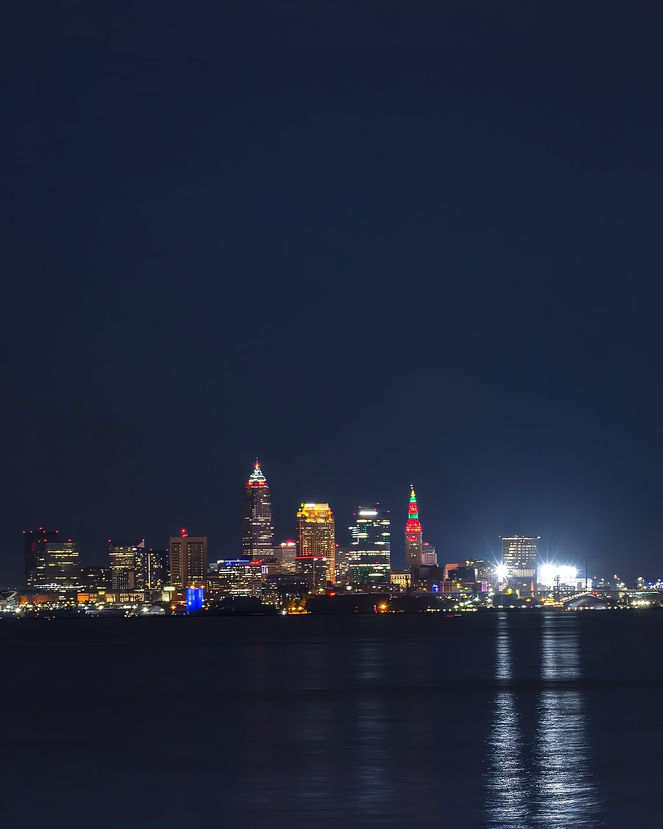 Nighttime city skyline with illuminated skyscrapers reflecting on calm water, including a tall building lit in red and green lights among other colorful lights.