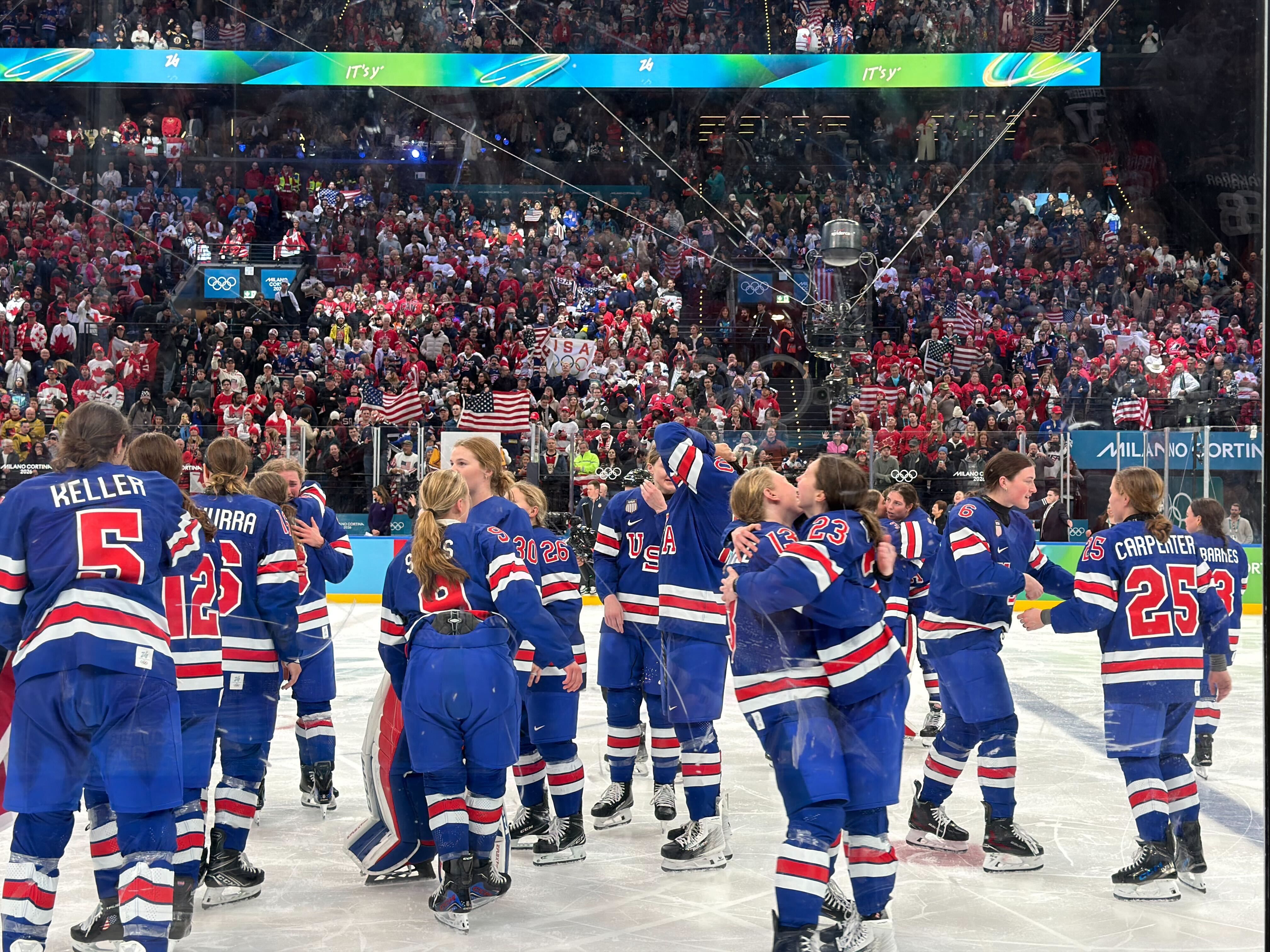 Female ice hockey players in blue USA uniforms celebrating on ice rink with cheering crowd waving American flags in background during Olympians event.