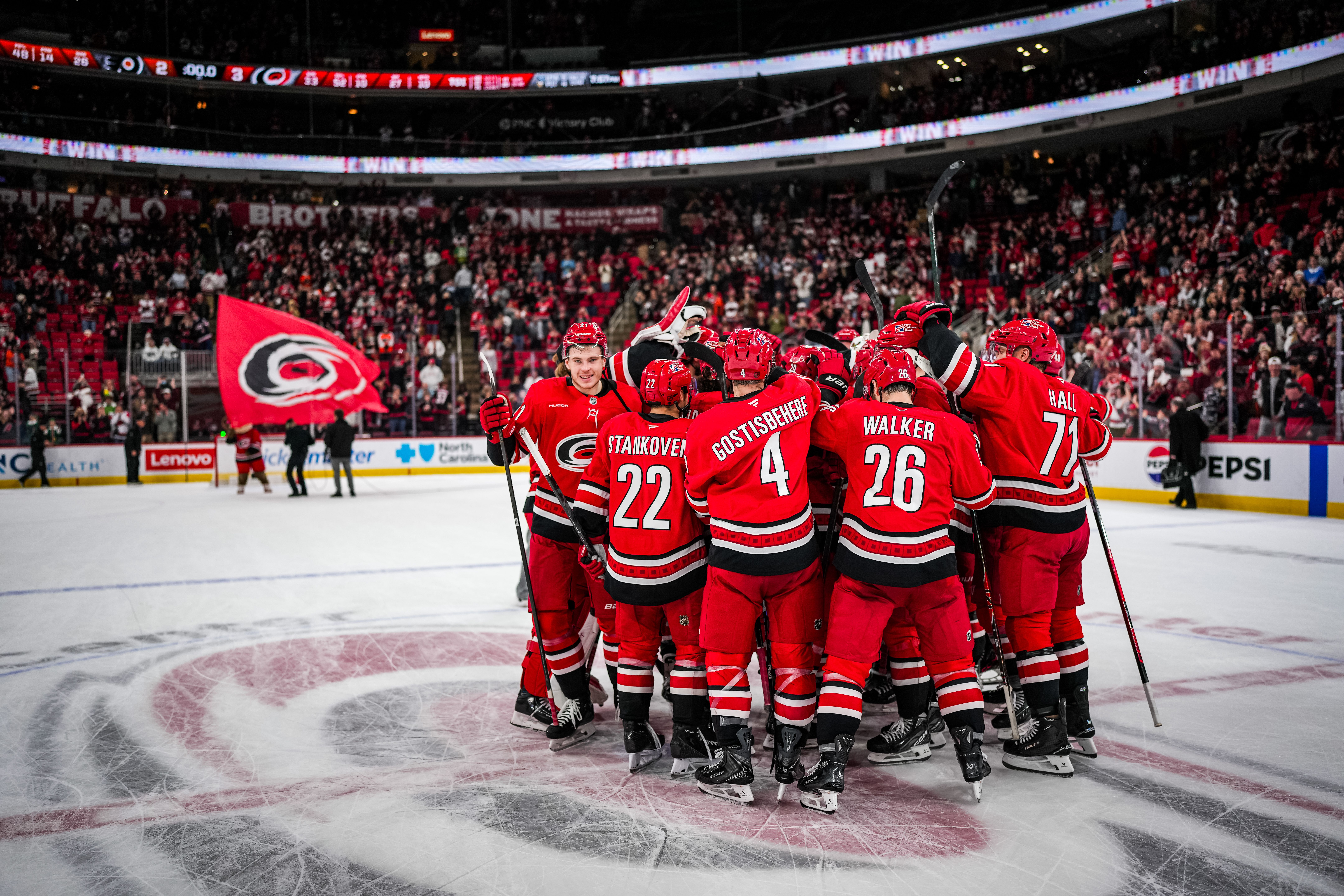 The Carolina Hurricanes celebrate a win by huddling at center ice as skaters in the background raise the team flag.
