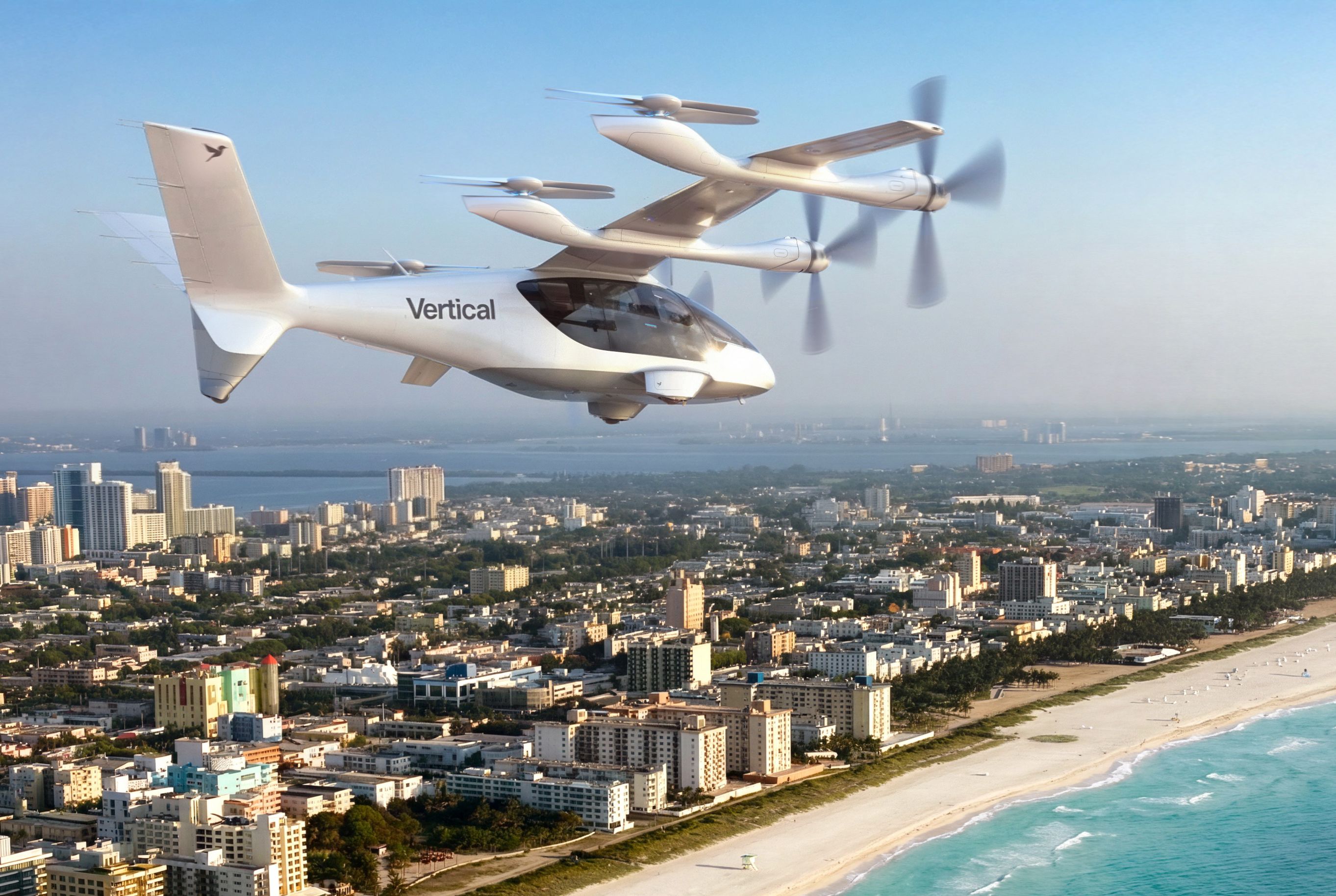 White tilt-rotor aircraft labeled "Vertical" flies over a coastal city with high-rise buildings, a sandy beach, and blue ocean under a clear sky.