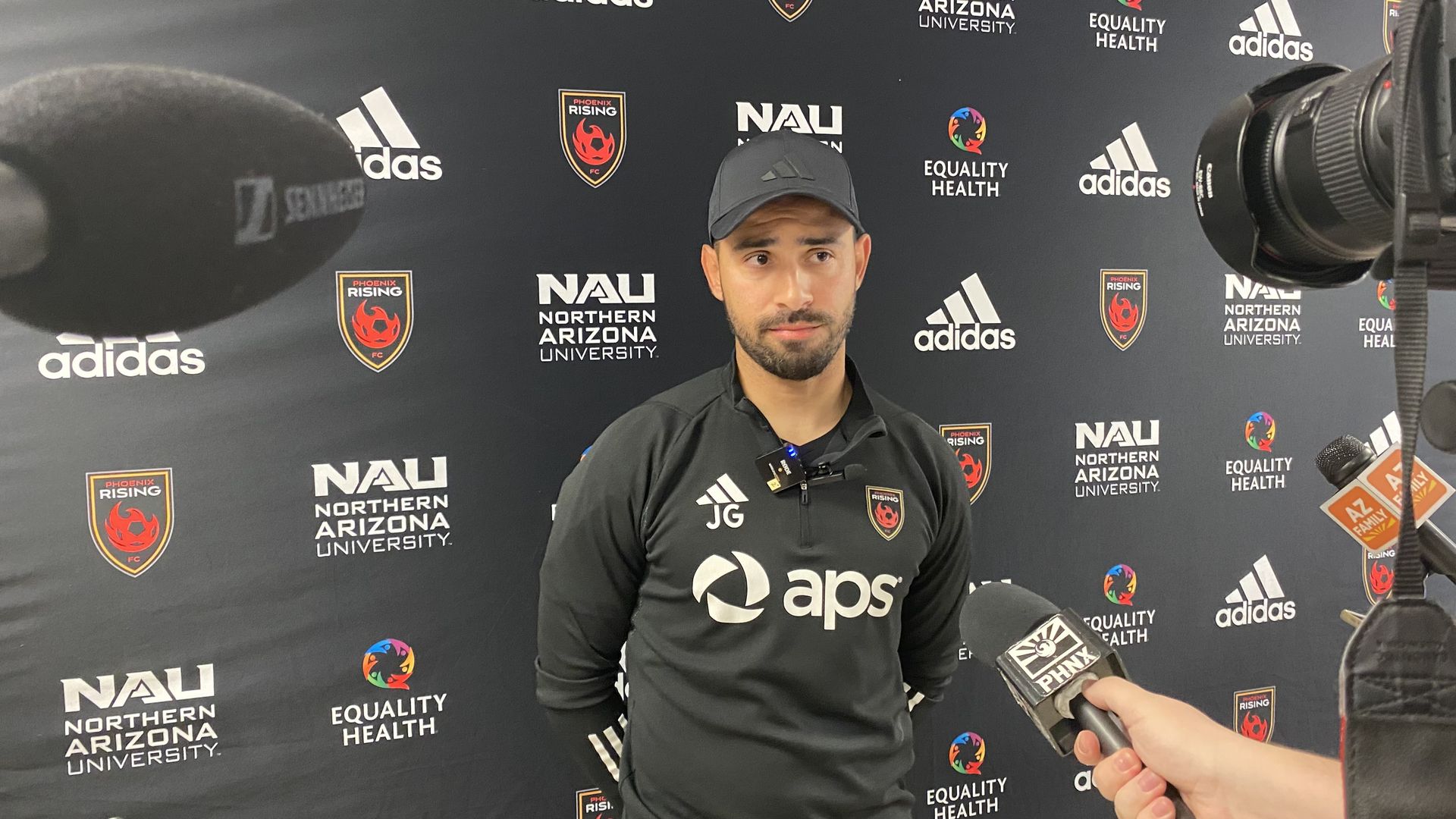 A man in a soccer jersey and a ballcap speaks to reporters.