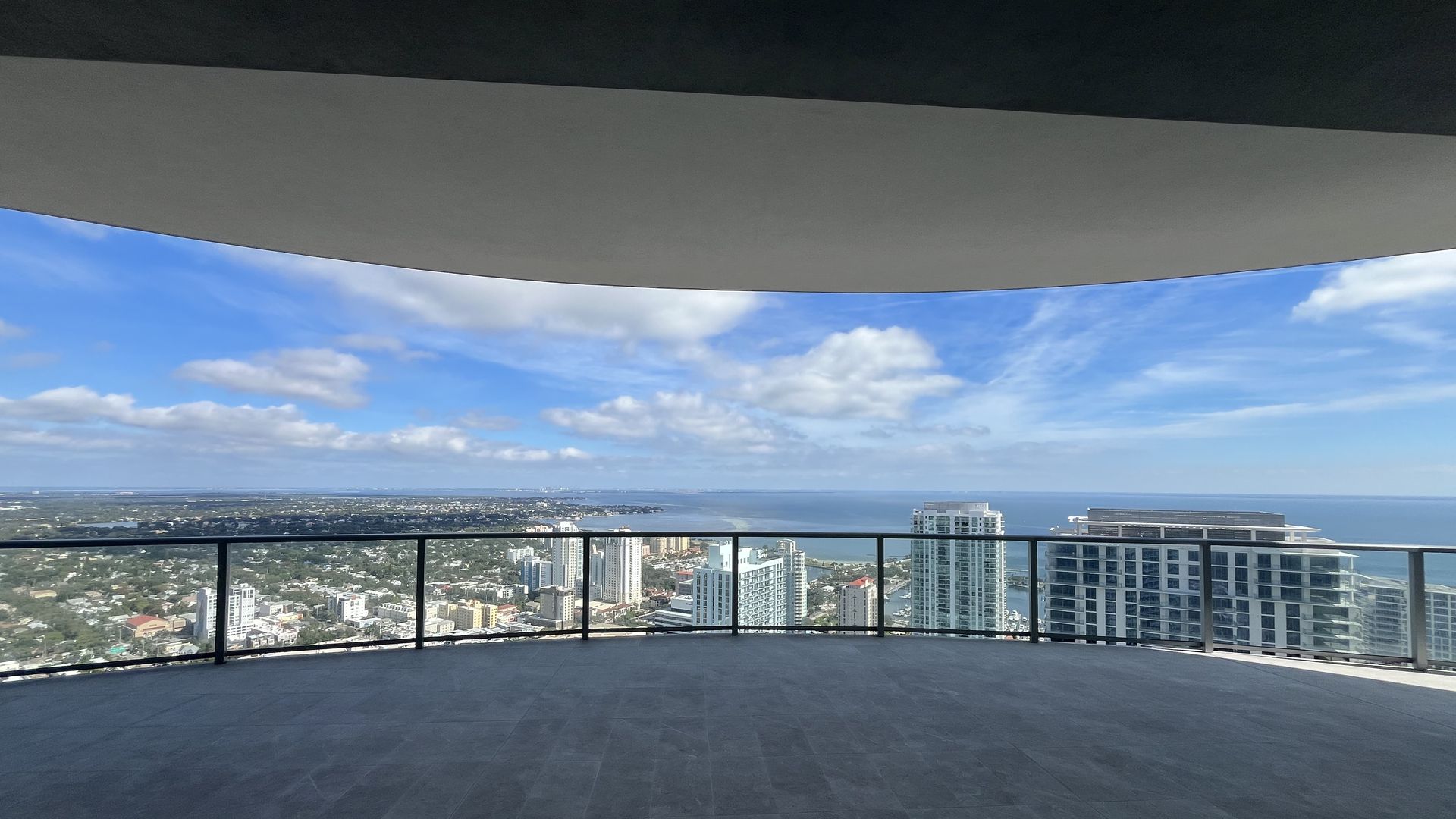Spacious curved balcony with glass railing overlooks city skyline, ocean, and blue sky with scattered clouds during daytime.