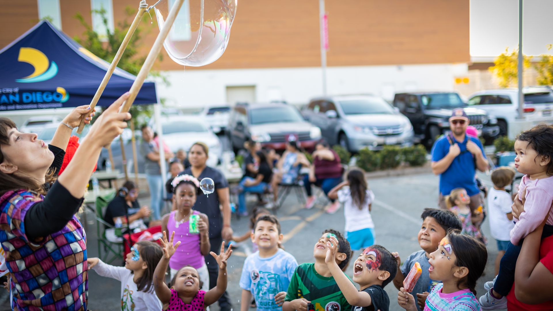 Children with ice cream and painted faces wearing colorful outfits reach toward large bubbles blown during a community block party, with a blue tent, parents, and parked cars in the background.