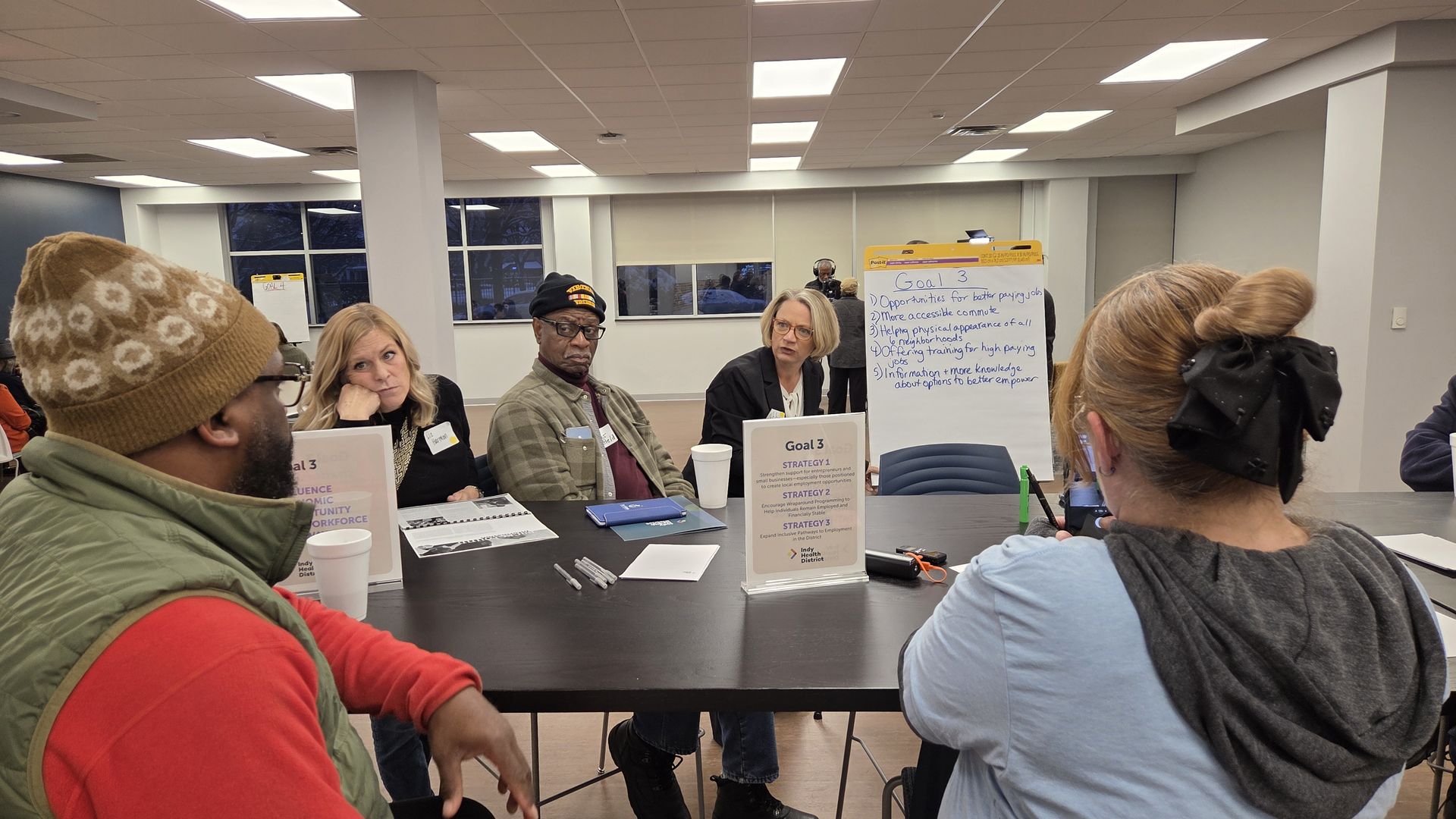 Five diverse people sit around a table in a meeting room, discussing goals written on a flipchart. One woman speaks while others listen attentively. The room has white walls and windows.