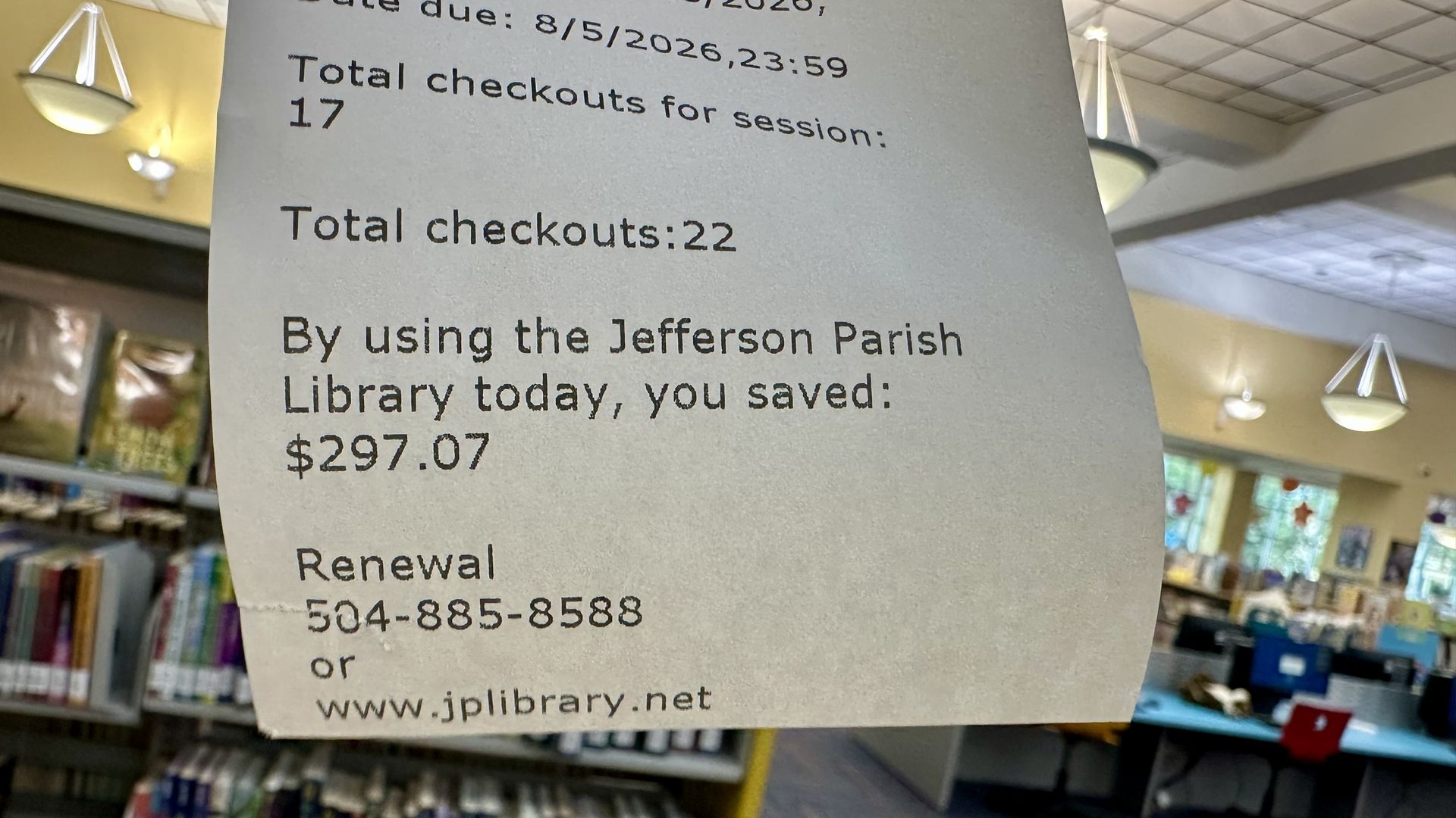 Gray library sign with black text showing checkout totals and savings: "Total checkouts for session: 17" and "Total checkouts: 22"; "you saved: $297.07". Background includes shelves and ceiling lights.