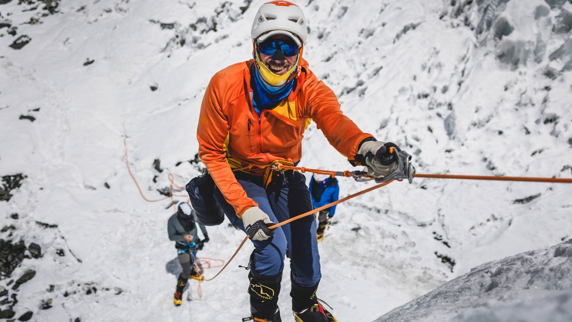A climber in an orange jacket and white helmet ascends a snowy mountain on a rope, wearing blue pants and protective gear, with other climbers in the background.