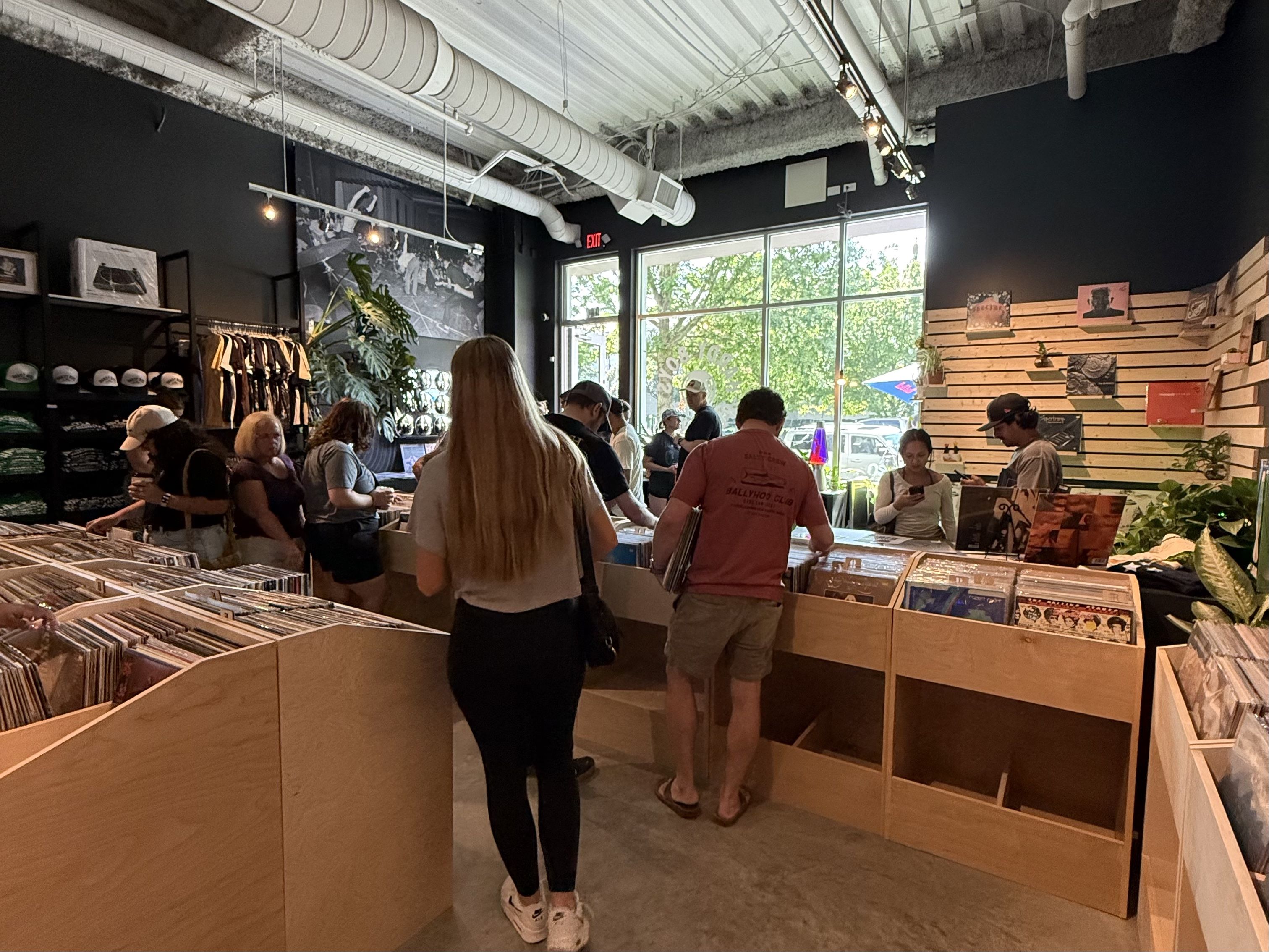Interior of a record store with people browsing vinyl records in wooden bins, black walls, large windows showing greenery outside, and shelves with hats and shirts on the left.