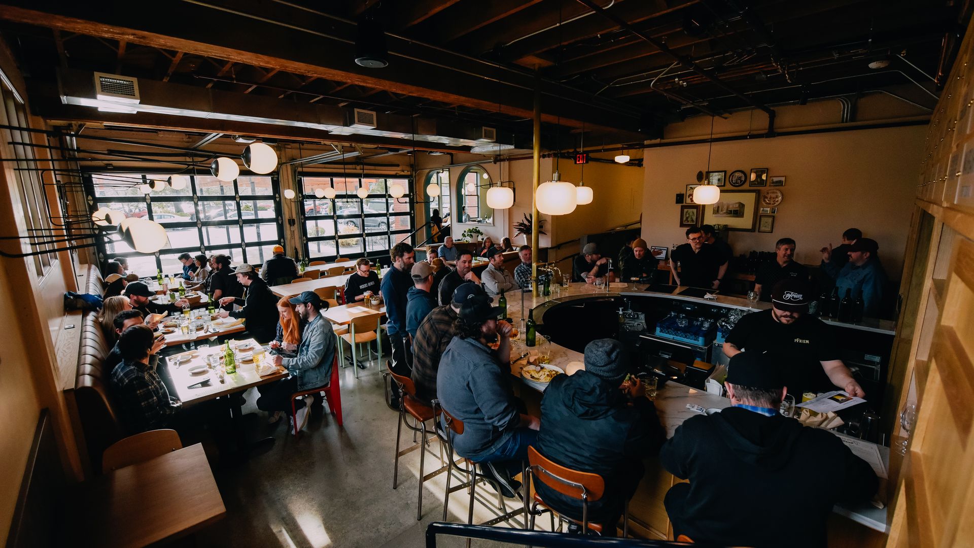 A wide shot shows a bunch of people in a brewery taproom sitting around a u-shaped bar and at small tables.