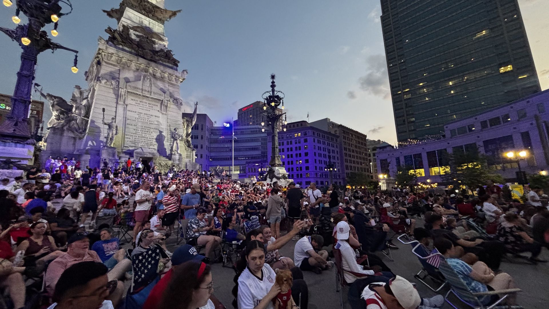 A large crowd sits and mingles around a white stone monument with statues in a city square at dusk. Purple lighting, ornate lampposts, and a tall dark office tower loom in the background.