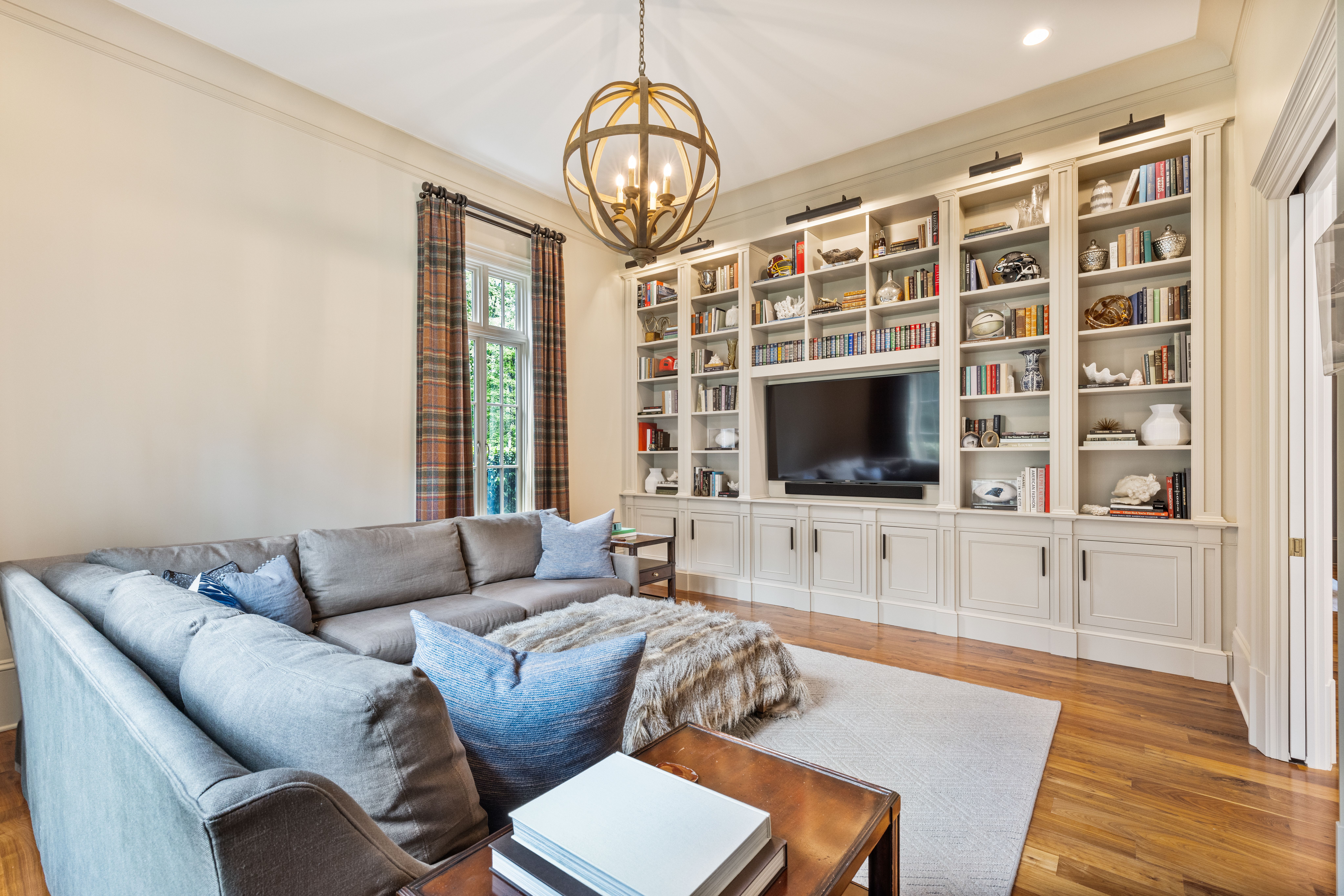 Bright living room with gray sectional sofa, blue pillows, furry ottoman, large window with plaid curtains, wooden floor, built-in white shelves filled with books and decor, and a round chandelier.